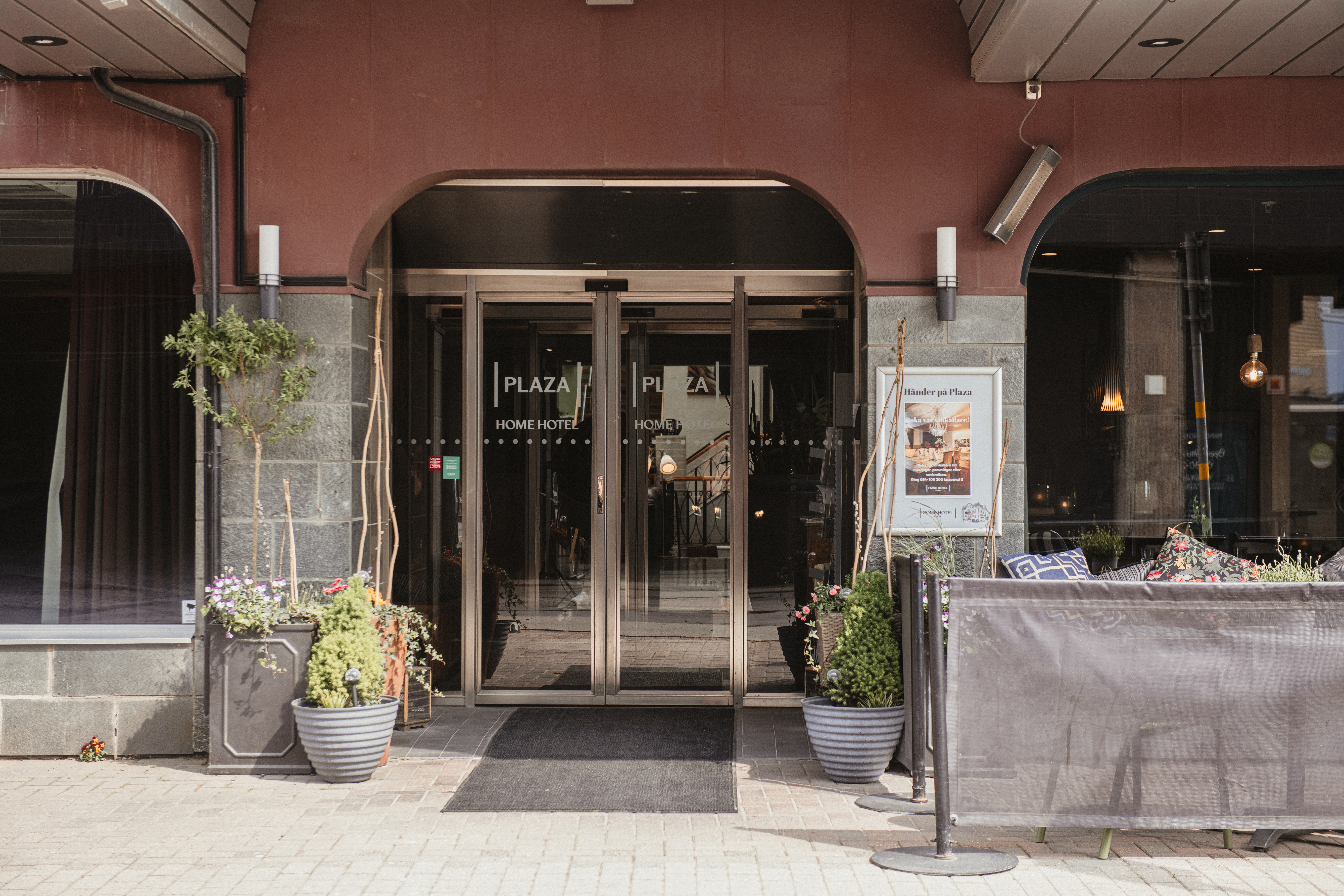 Ingången av ett hotell med glasdörrar och krukväxter, omgiven av en röd och grå fasad. Text på dörrarna: "PLAZA HOME HOTEL".