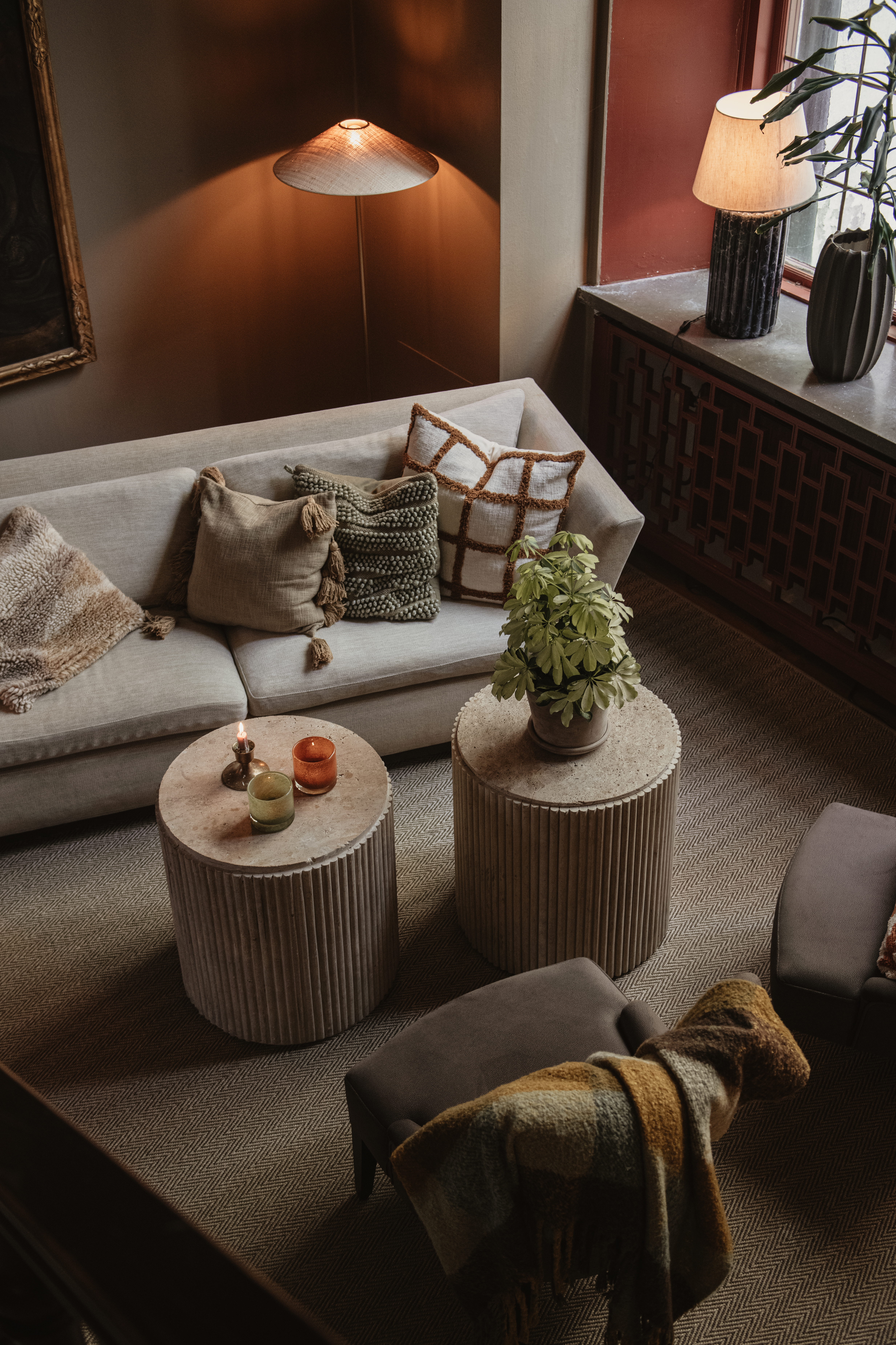En beige sofa med dekorative puder står i en hyggelig stue. To runde borde har lys og en plante. En varm lampe og et vindue oplyser rummet.