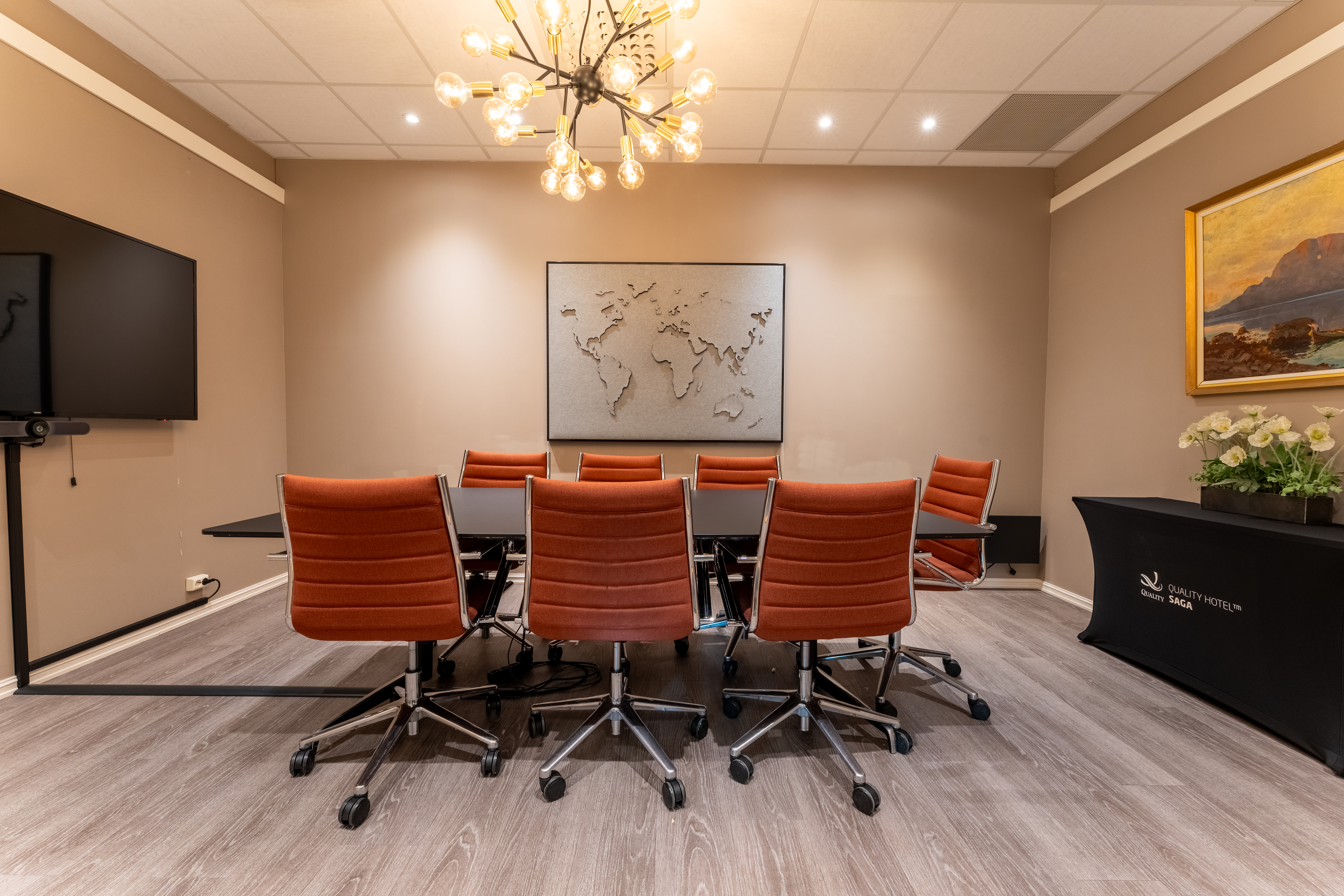 Hyvin valaistu kokoushuone, jossa on pitkä pöytä, oranssit tuolit, suuri näyttö ja maailmankartta. Telineessä lukee Quality Hotel Saga.