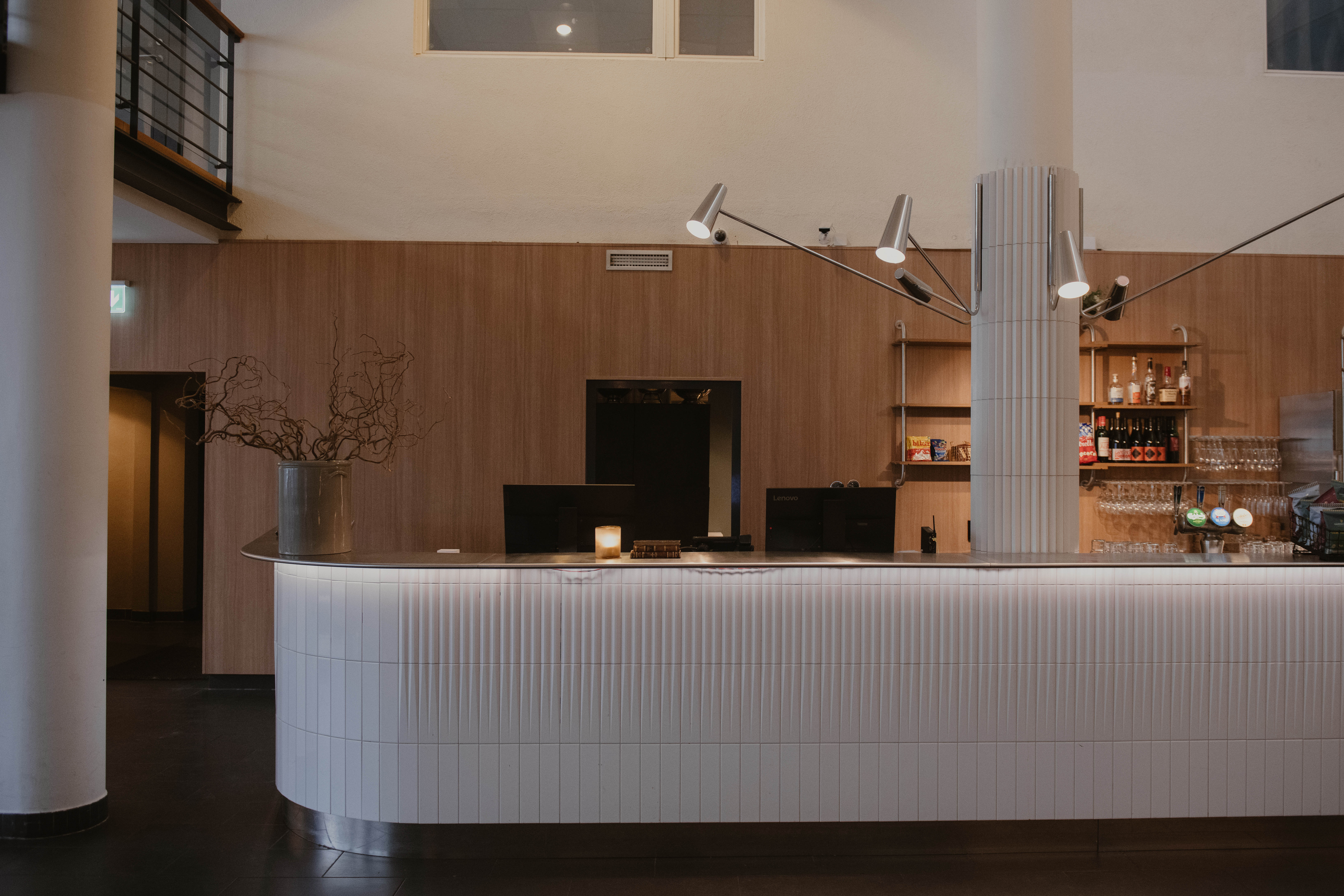 En modern receptionsdisk med rundad design står i en varmt belyst lobby. Bakom den, trähyllor visar flaskor, och en elegant belysning hänger ovanför.