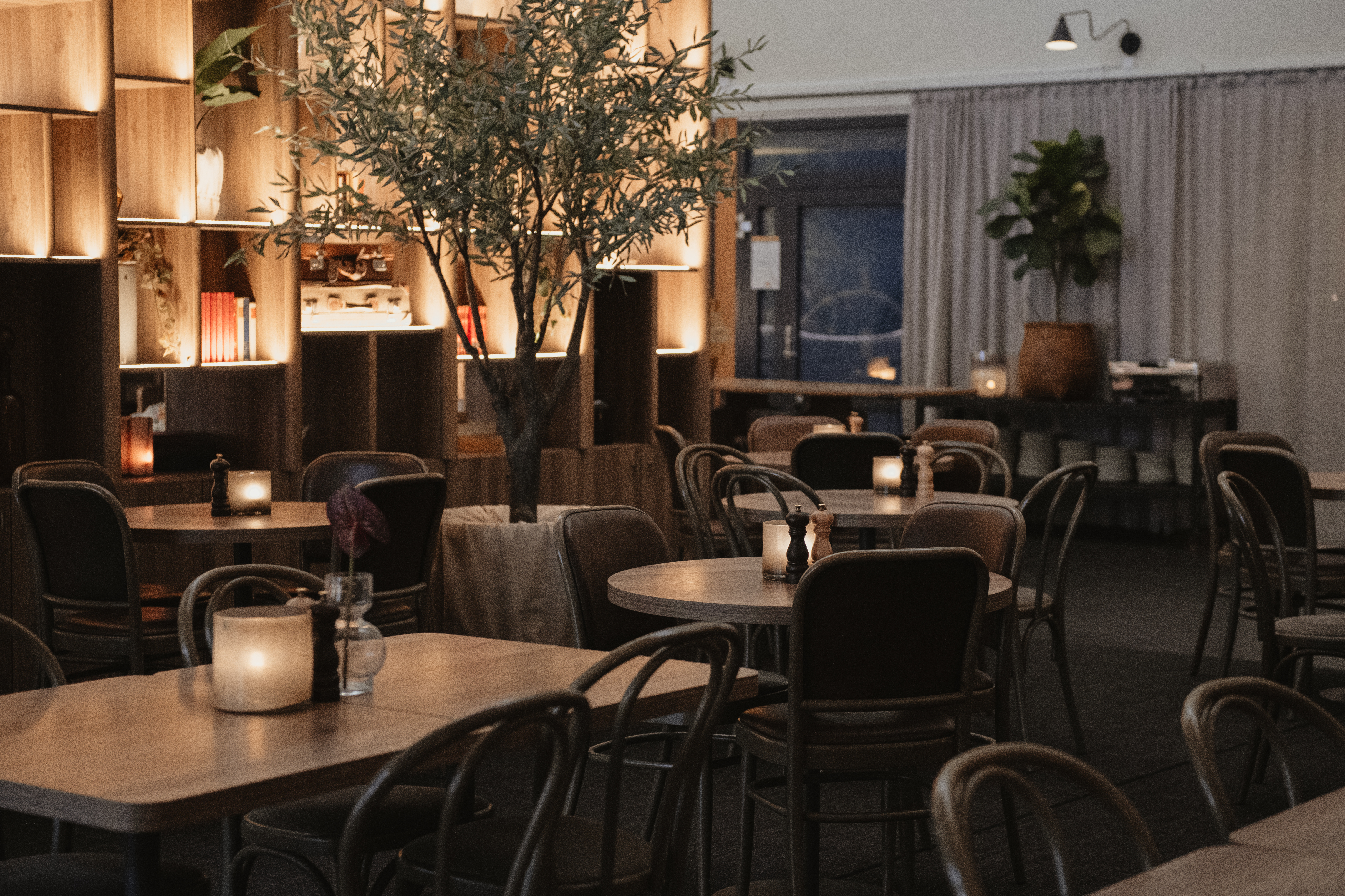 Bord med trästolar är arrangerade i en svagt upplyst restaurang med mjuka ljus, växter och ljus. En stor hylla med dekorativa föremål ger en varm och mysig atmosfär.