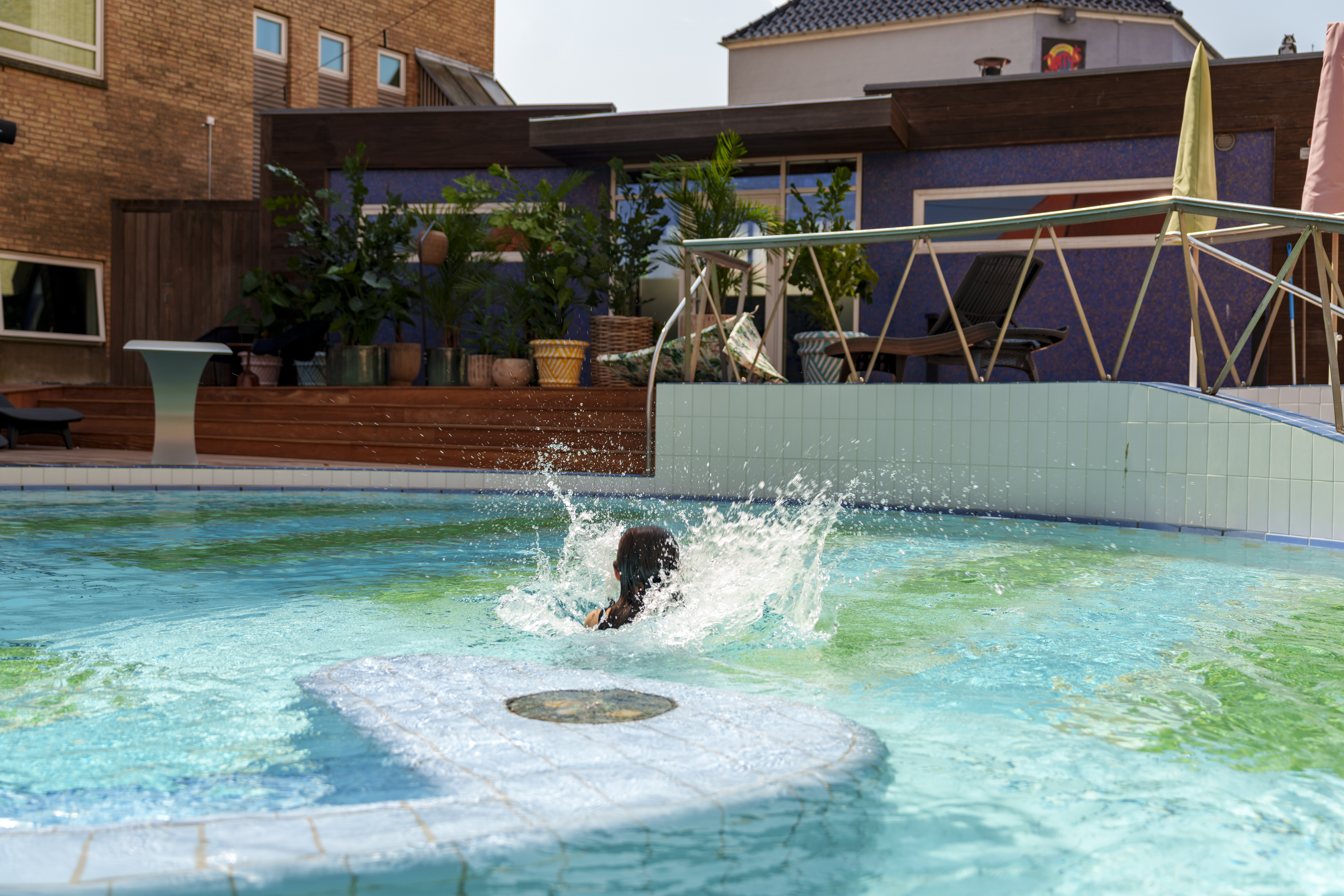 En person plasker i en blå pool; omkring dem er potteplanter og moderne bygninger i urbane omgivelser.