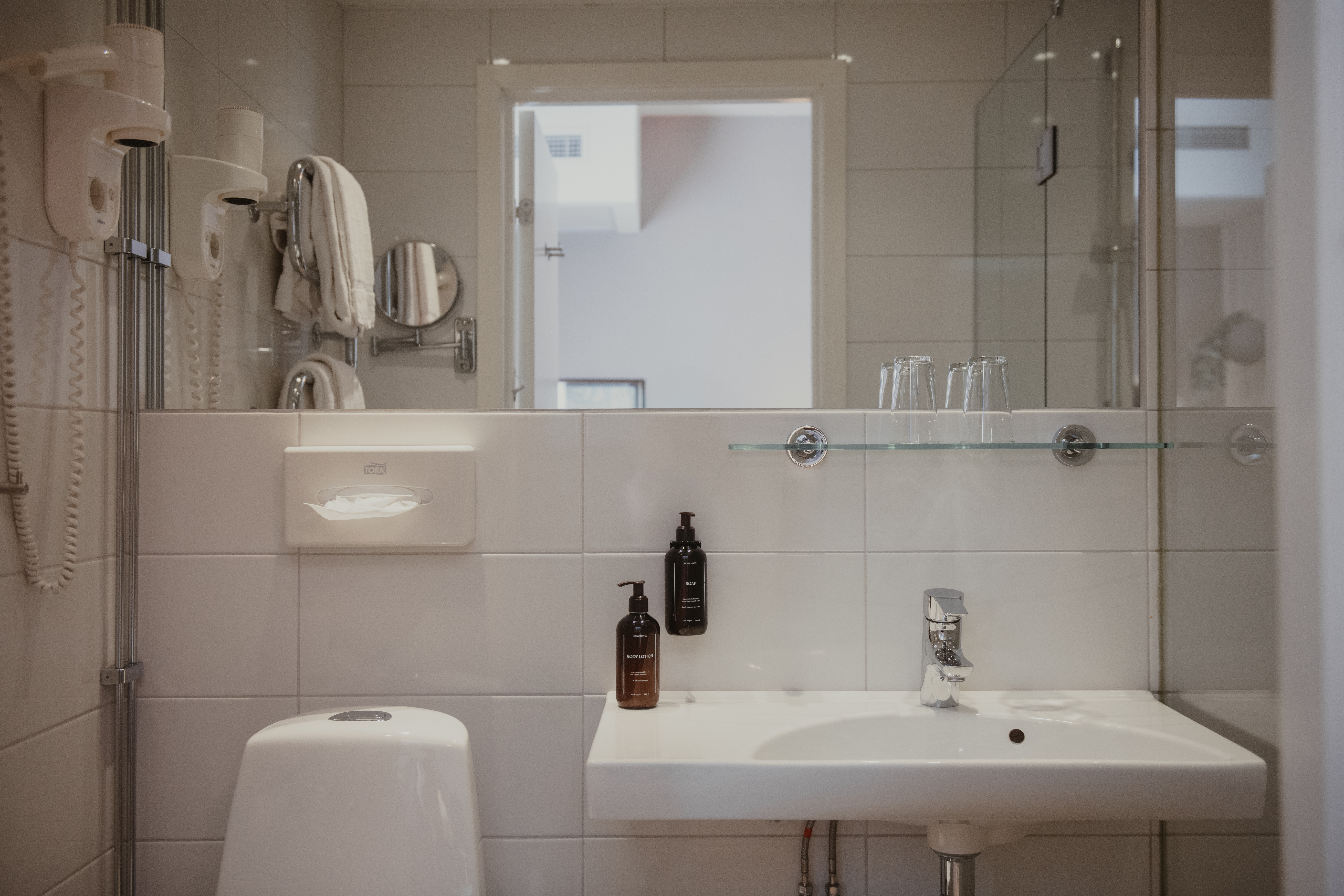 Handfat med kran i ett vitkaklat badrum; flankerad av tvåltvålpumpar och glas. I närheten finns en toalett, väggmonterad hårtork och handdukshängare som förstärker hotellets enkla, funktionella estetik.