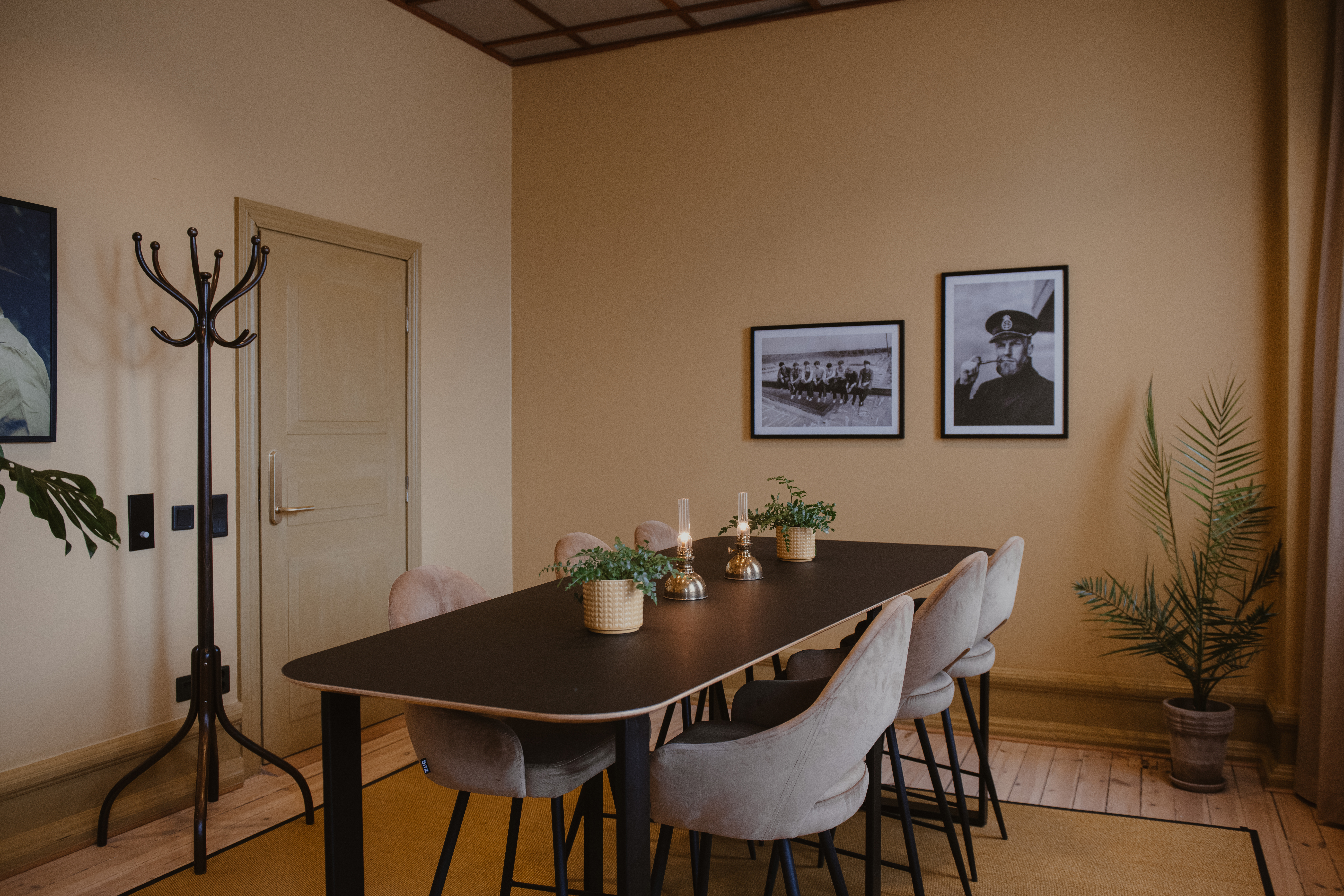 Spisebord med bløde stole er dekoreret med potteplanter og stearinlys, placeret i et varmt oplyst rum med beige vægge, indrammede fotografier og en stumtjener i nærheden.