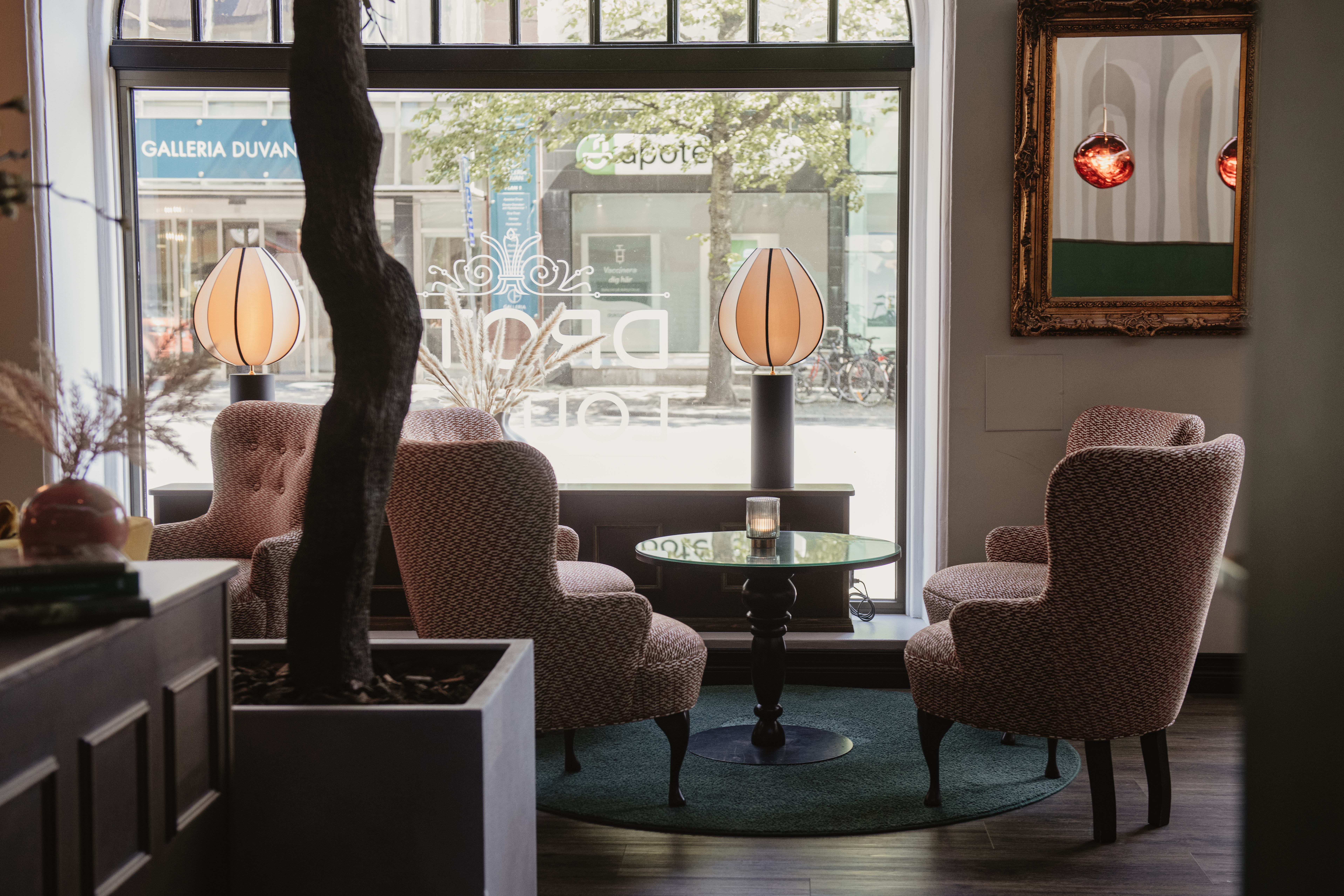 Fyra stoppade fåtöljer omger ett litet runt bord på en grön matta i ett mjukt upplyst café. Stora fönster visar utomhusskyltar: "APOTEK" och "GALLERIA DUVAN", med träd som syns utanför.
