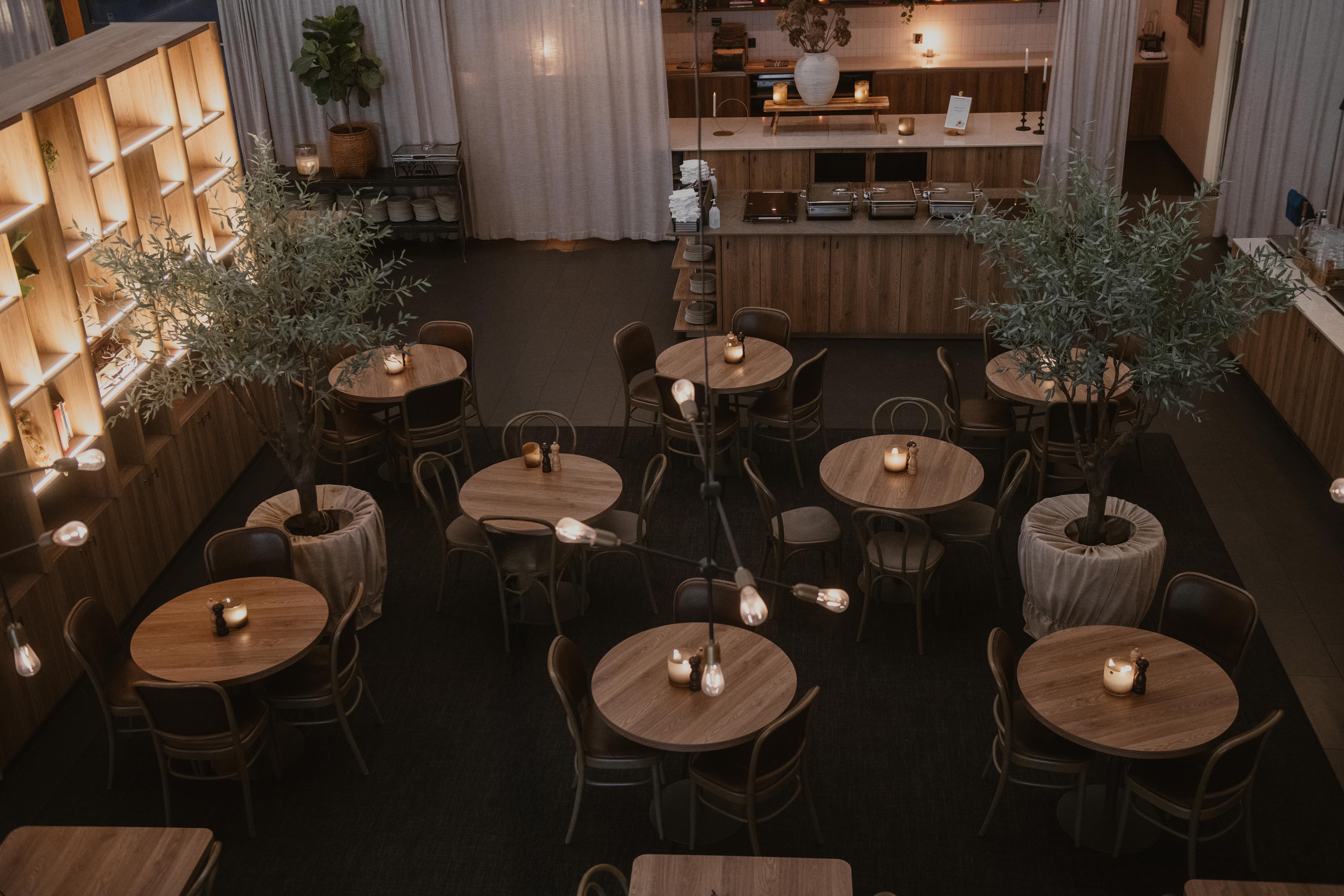 Runda träbord med tända ljus arrangerade i en mysig, svagt upplyst restaurang med krukträd och mjuka gardiner. Området runt omkring inkluderar en disk och hyllor med dekorativa föremål.