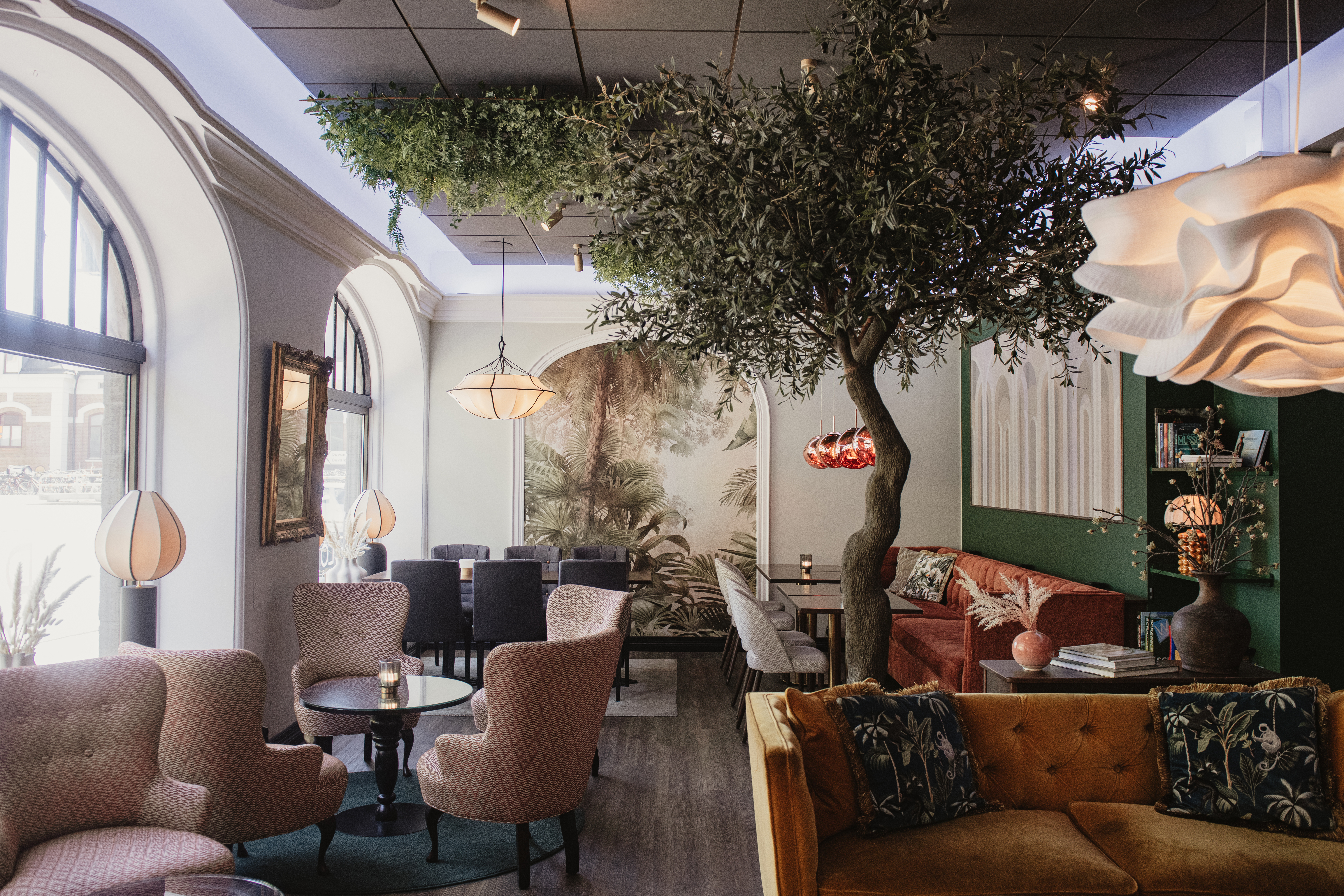 Ett träd dekorerar ett stiligt, mysigt café med bekväma sittplatser, eleganta lampor och stora välvda fönster som släpper in naturligt ljus. En muralmålning som föreställer naturen bidrar till den inbjudande atmosfären.