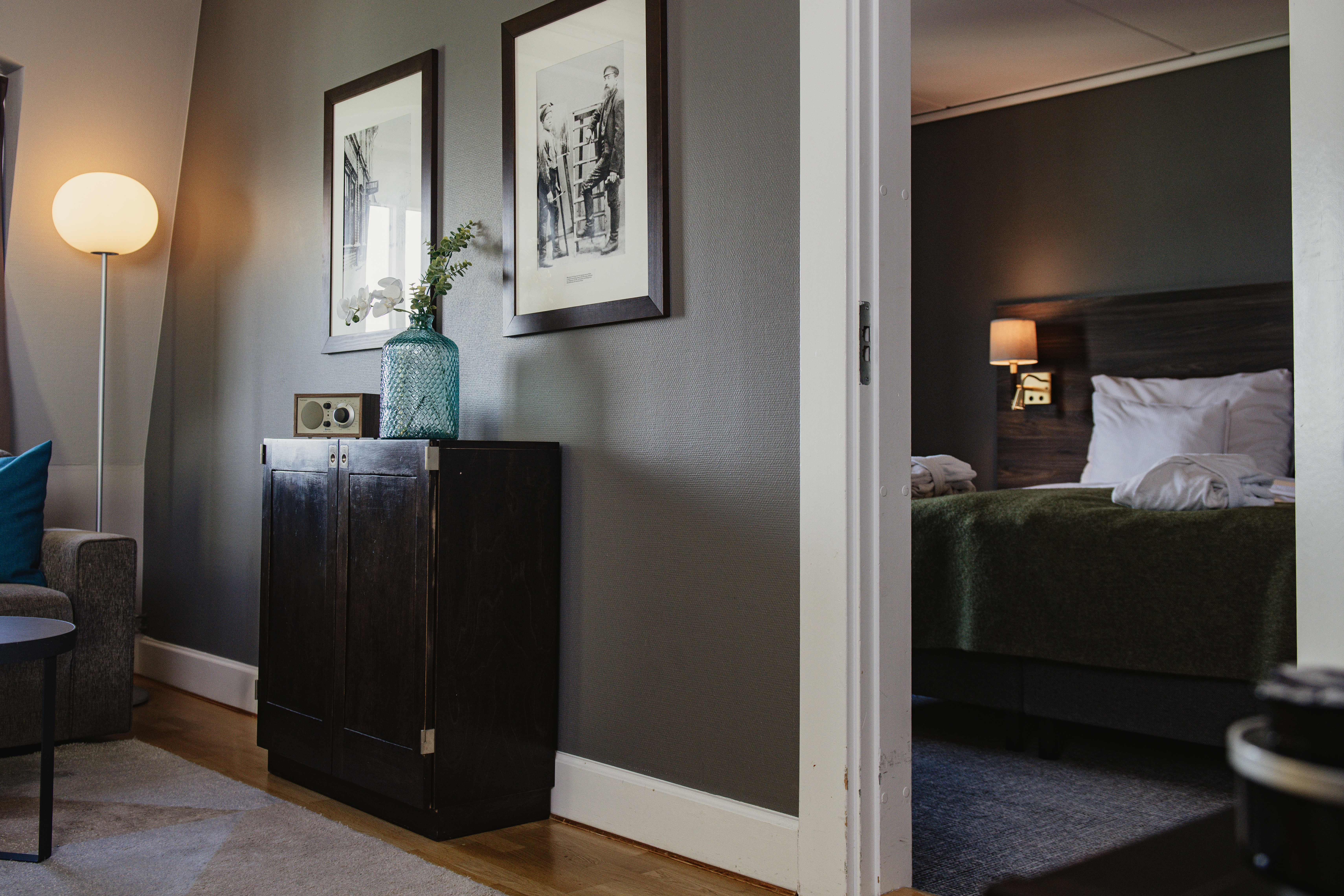 A dark wooden cabinet with a vase and clock sits next to a lit floor lamp; through an open doorway, a bedroom with a neatly made bed and wall lamp is visible.