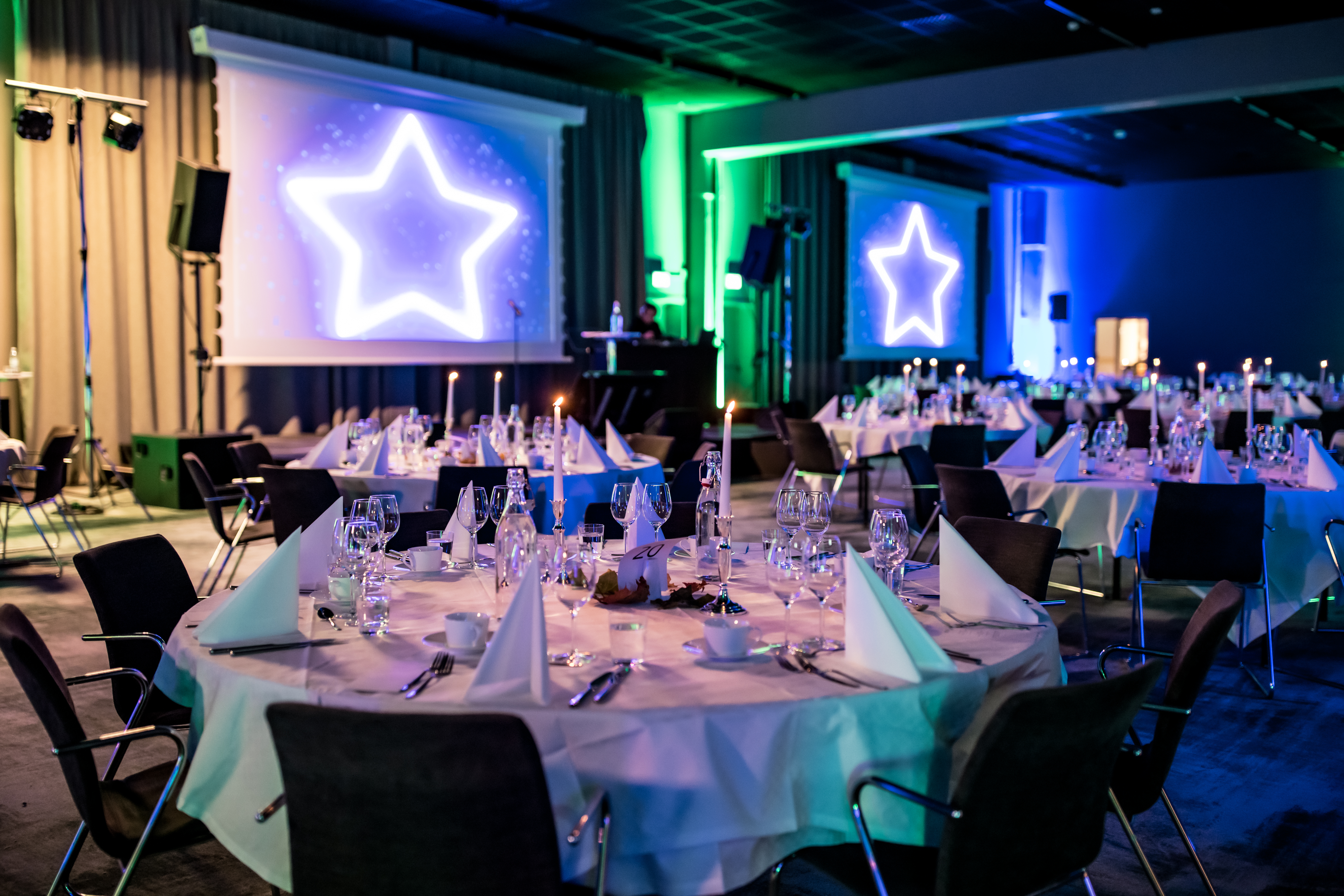 Et festlig arrangementslokale med runde bord dekket til middag, opplyst av fargerike lys og stjerneprojeksjoner.