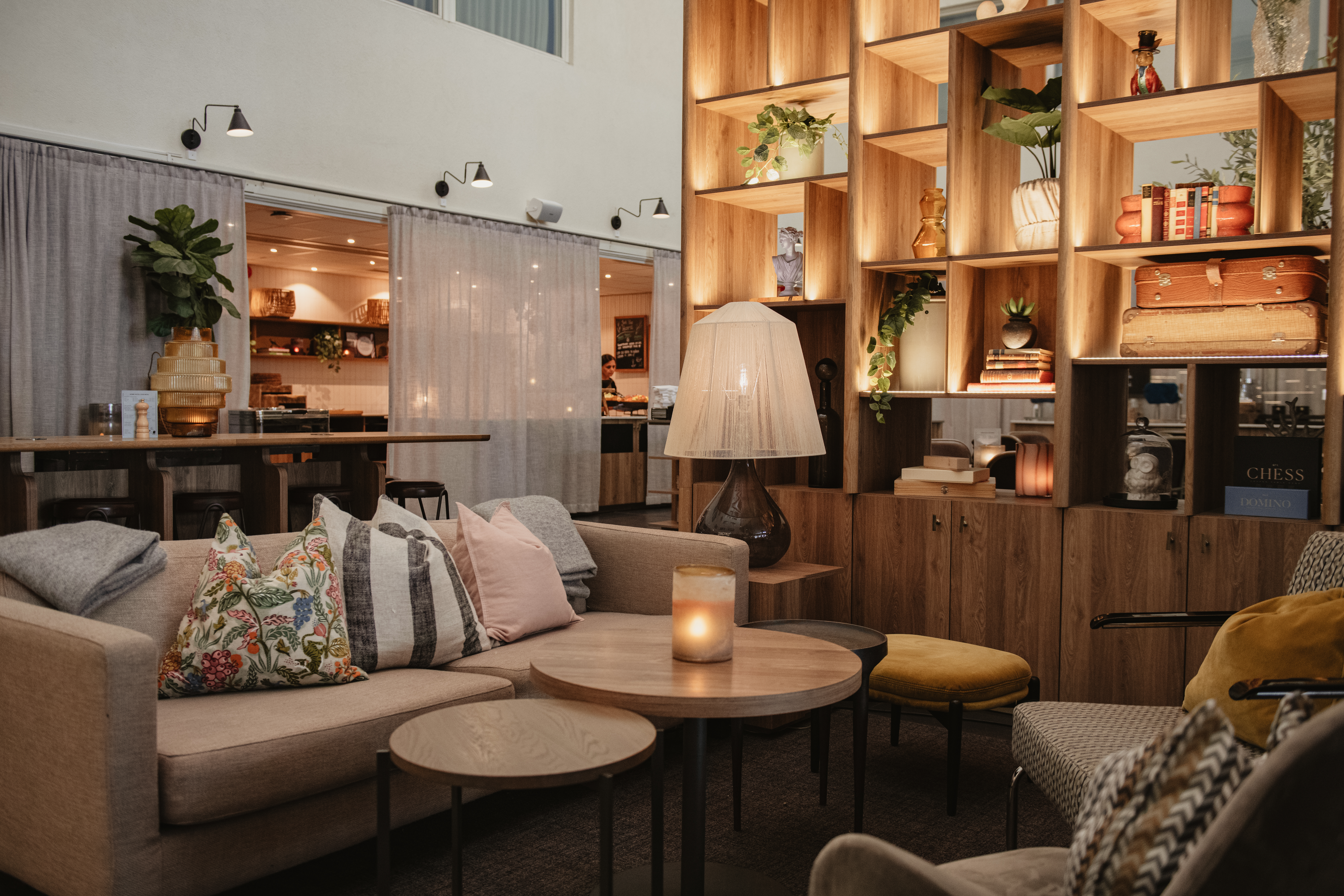 En mysig lounge har beige soffor med färgglada kuddar, ett runt träbord med ett tänt ljus och en hög hylla med böcker och växter i ett svagt upplyst rum.