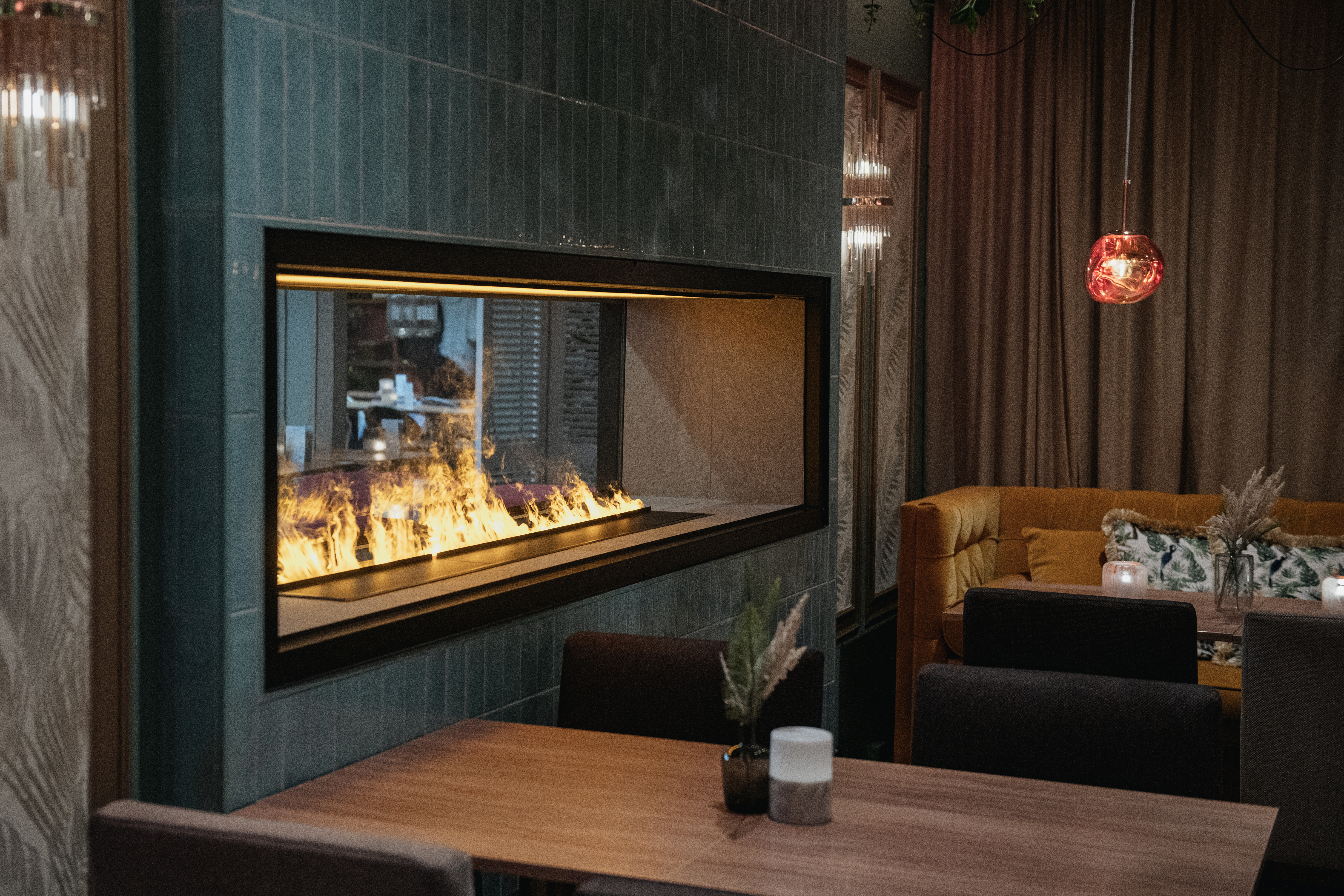En modern eldstad med flammor brinnande stadigt är inbyggd i en kaklad vägg. Omkring den finns bord, stolar och en mysig bås i en varmt belyst, stilren restaurang.