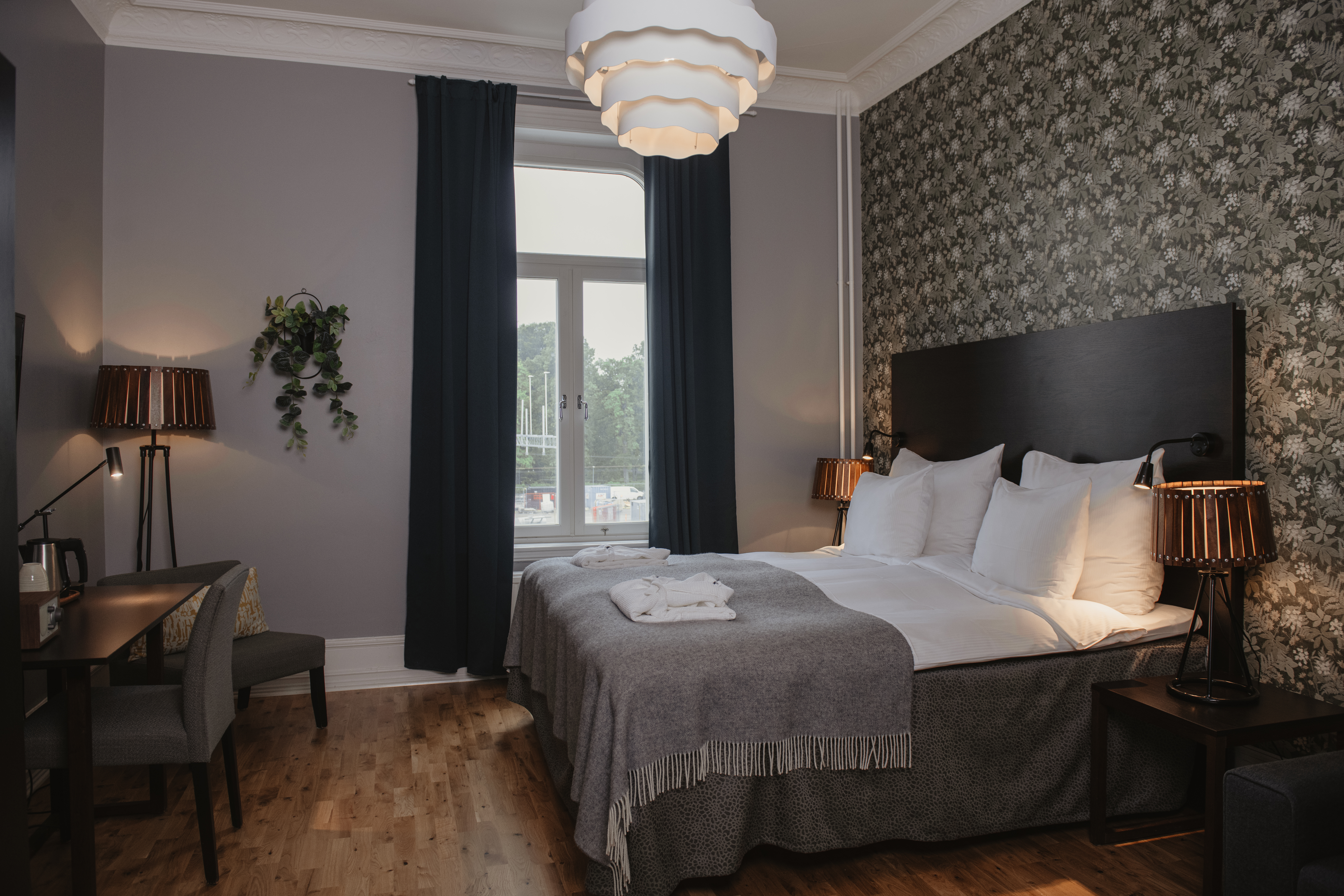 Mukava hotellihuone sisältää siististi pedatun sängyn valkoisin vuodevaattein kuvioitua seinää vasten. Kirjoituspöytä ja tuoli ovat lähellä pehmeässä valaistuksessa. Ikkunasta on näkymä ulos.