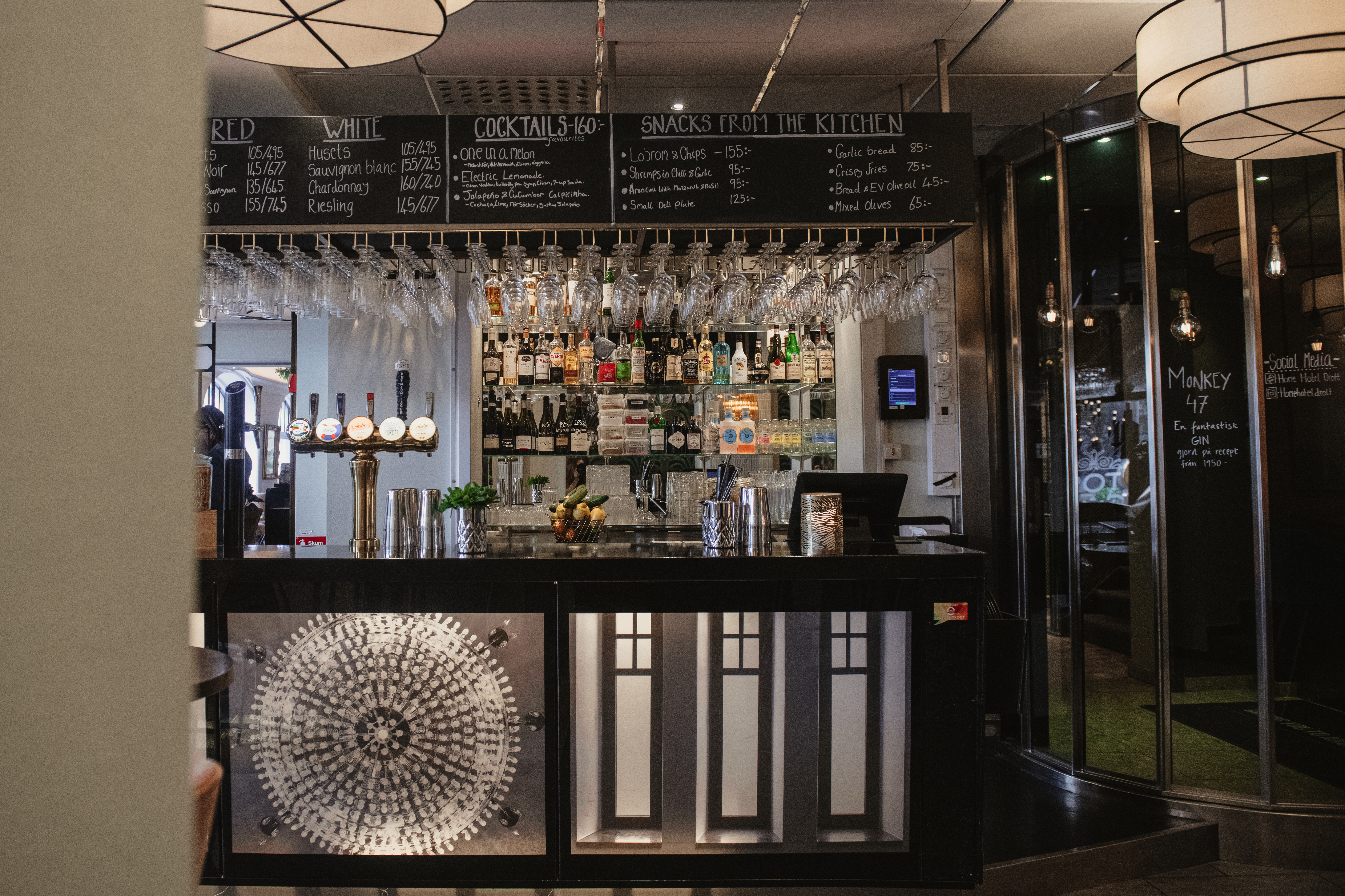 Bar med flaskor och glas uppradade, bakbelysta och organiserade. Svarta tavlan ovan listar drycker och snacks: "RED, WHITE, COCKTAILS 40 kr, SNACKS FROM THE KITCHEN." Elegant interiör med modern inredning.
