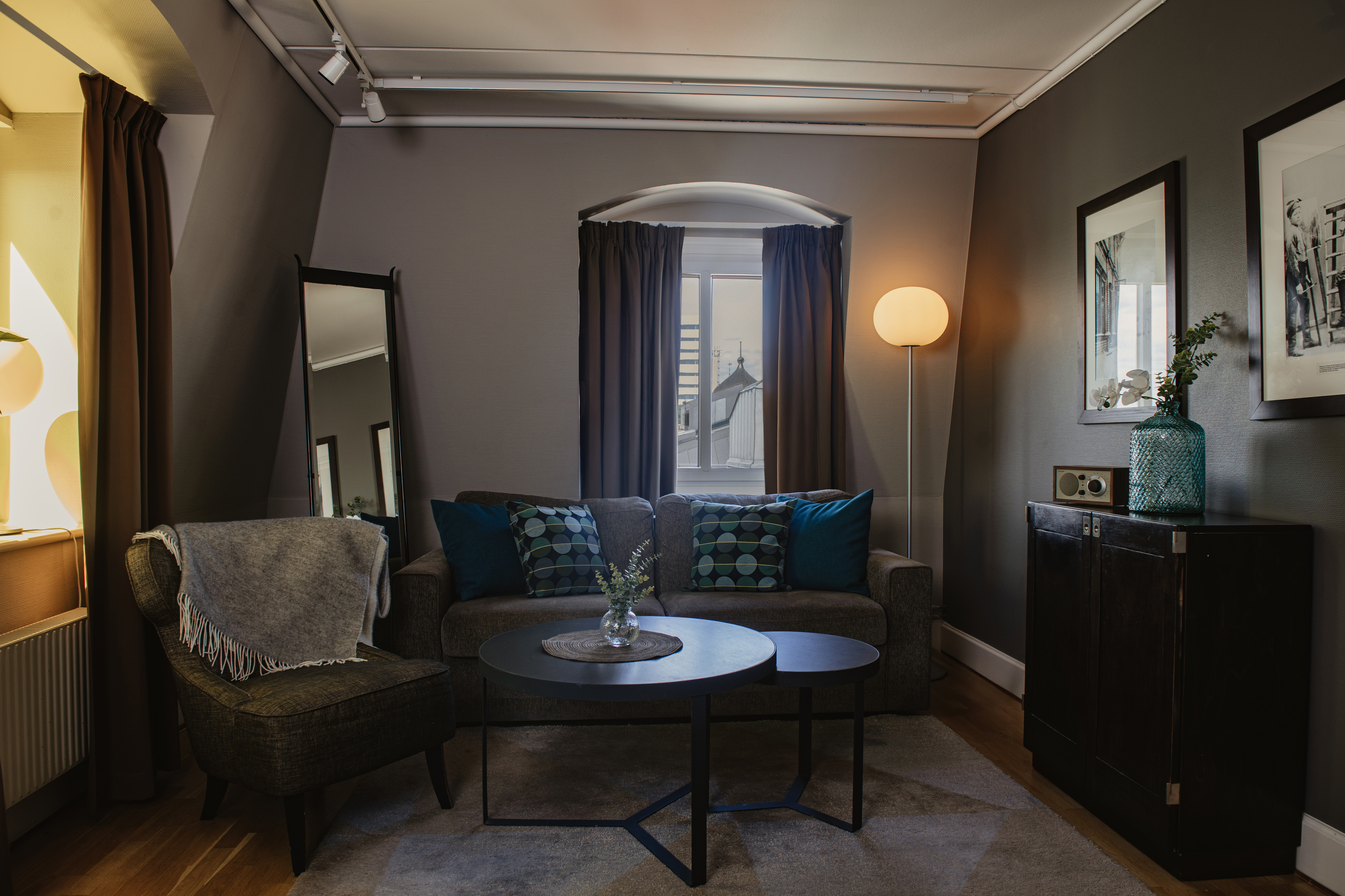 A cozy living room features a grey sofa with blue cushions and a side chair, complemented by a round coffee table. A floor lamp and framed pictures enhance the warm, inviting ambiance.