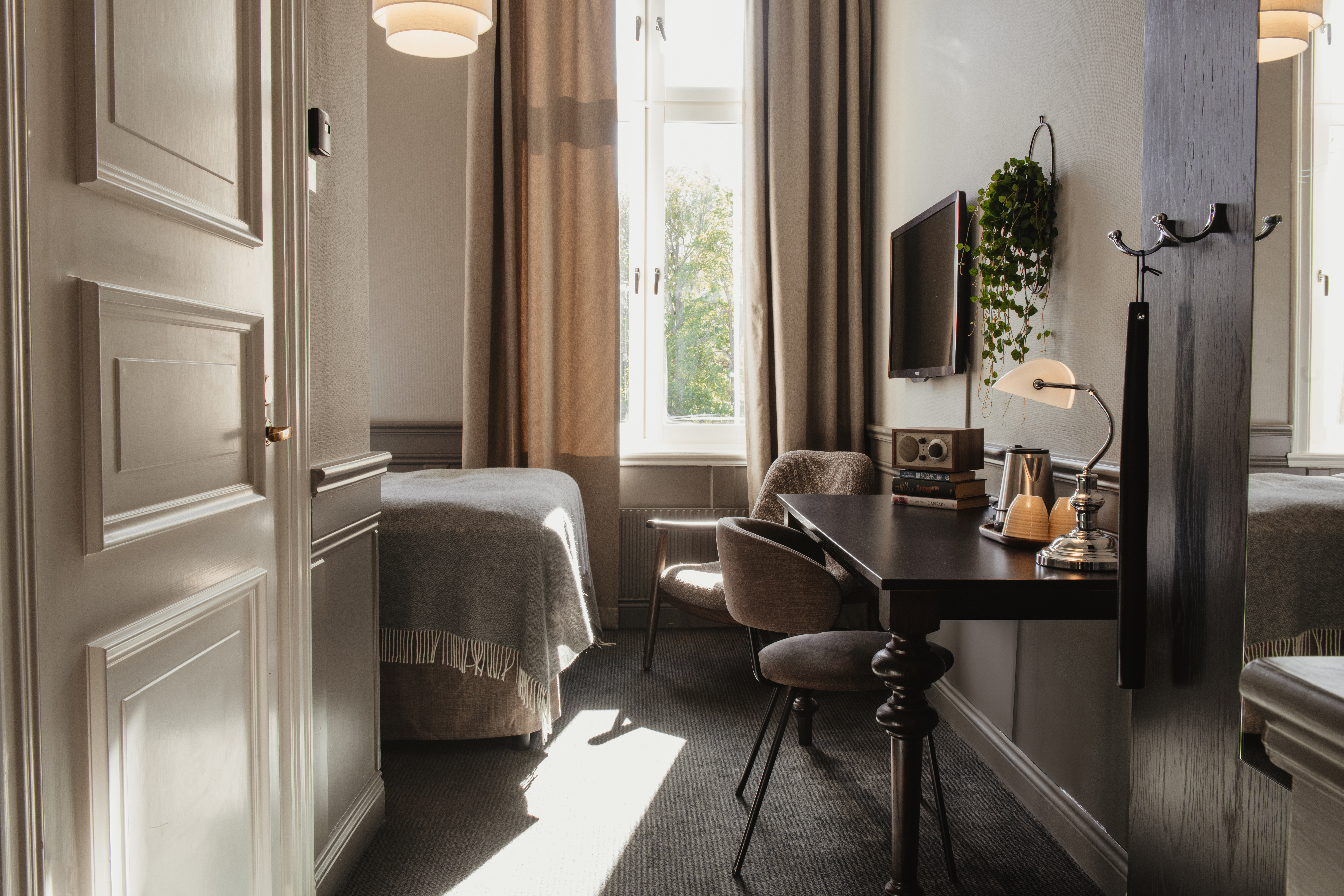 Ett varmt och inbjudande hotellrum badat i naturligt ljus, med en bekväm säng, en funktionell skrivbordsplats och fönsterutsikt.