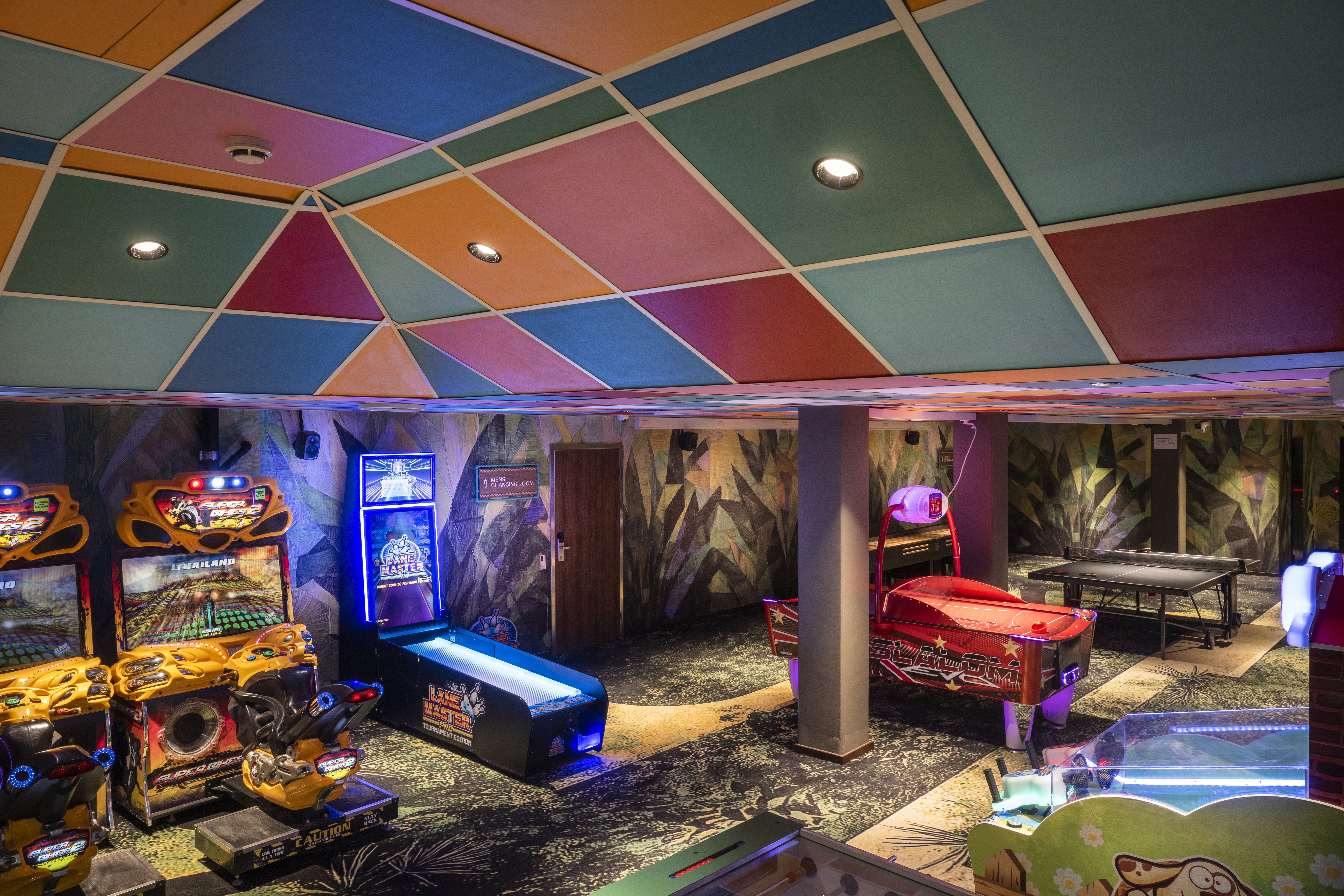 Arkadespil står i et farverigt rum, inklusive racerspil, airhockey og flipperspil, under et livligt, mønstret loft. Neonlys oplyser rummet og skaber en spændende spilatmosfære.