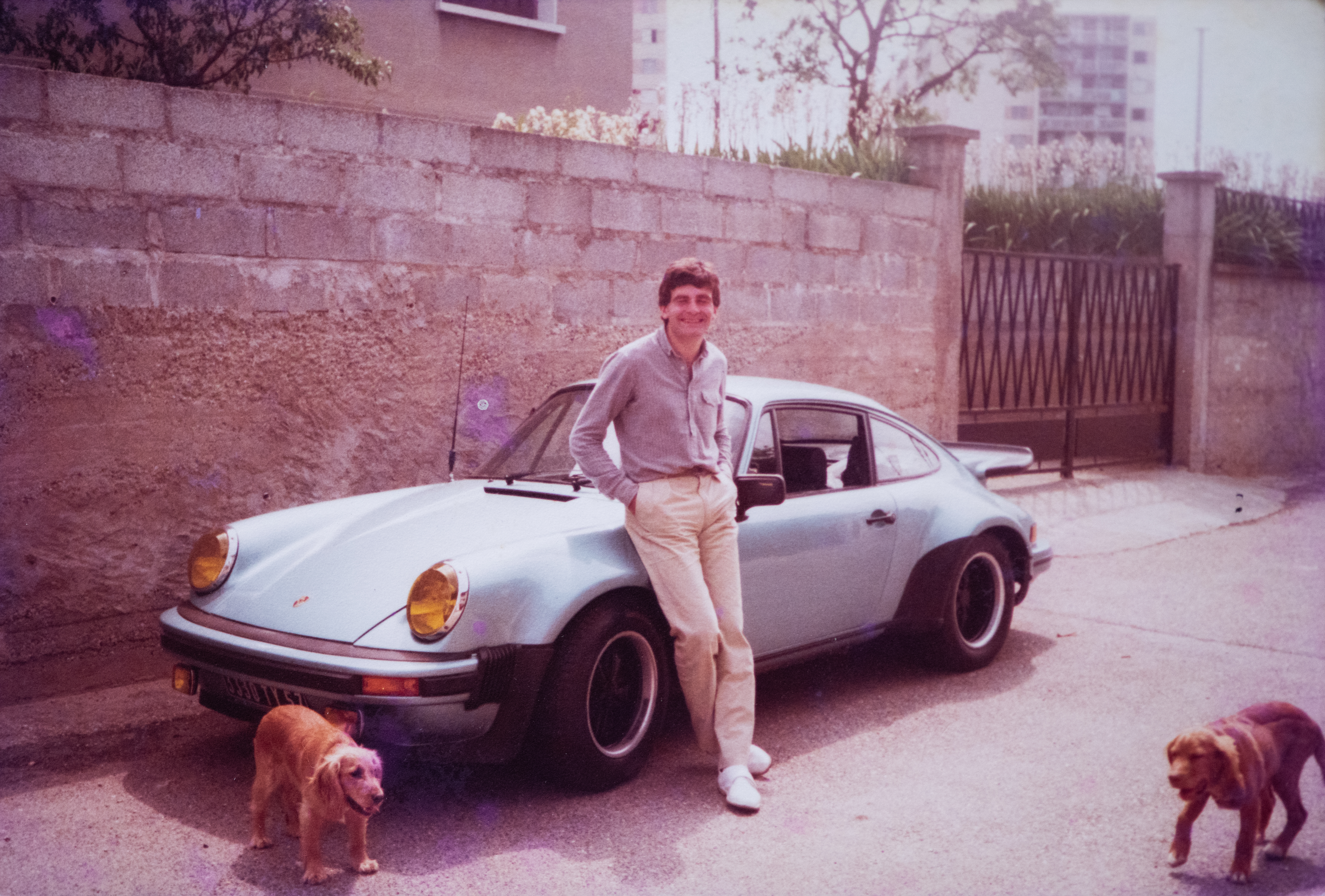 Old photo of man leaning against a blue Porsche. Dogs in foreground