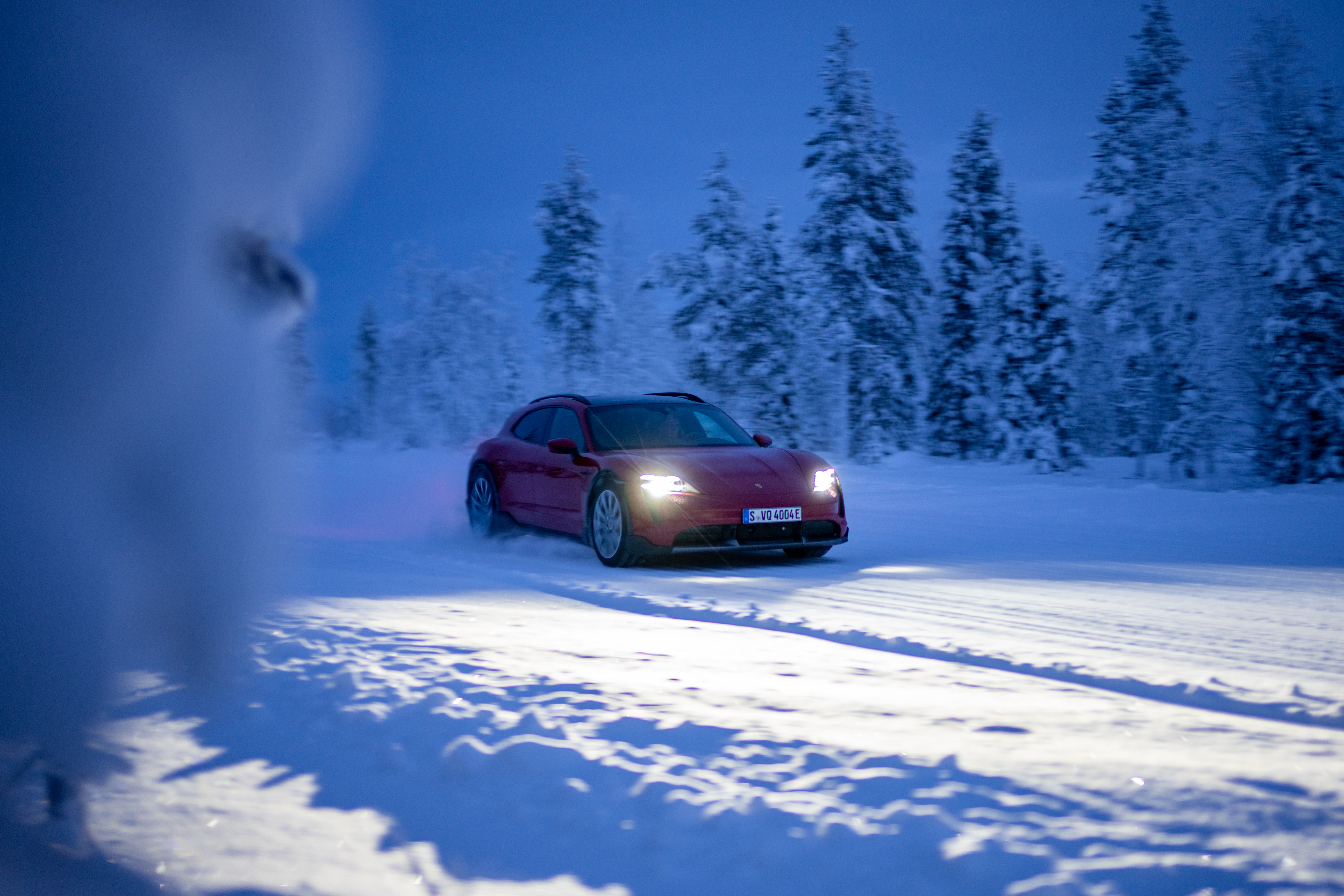 Red Porsche Taycan, headlights on, driving on snowy road