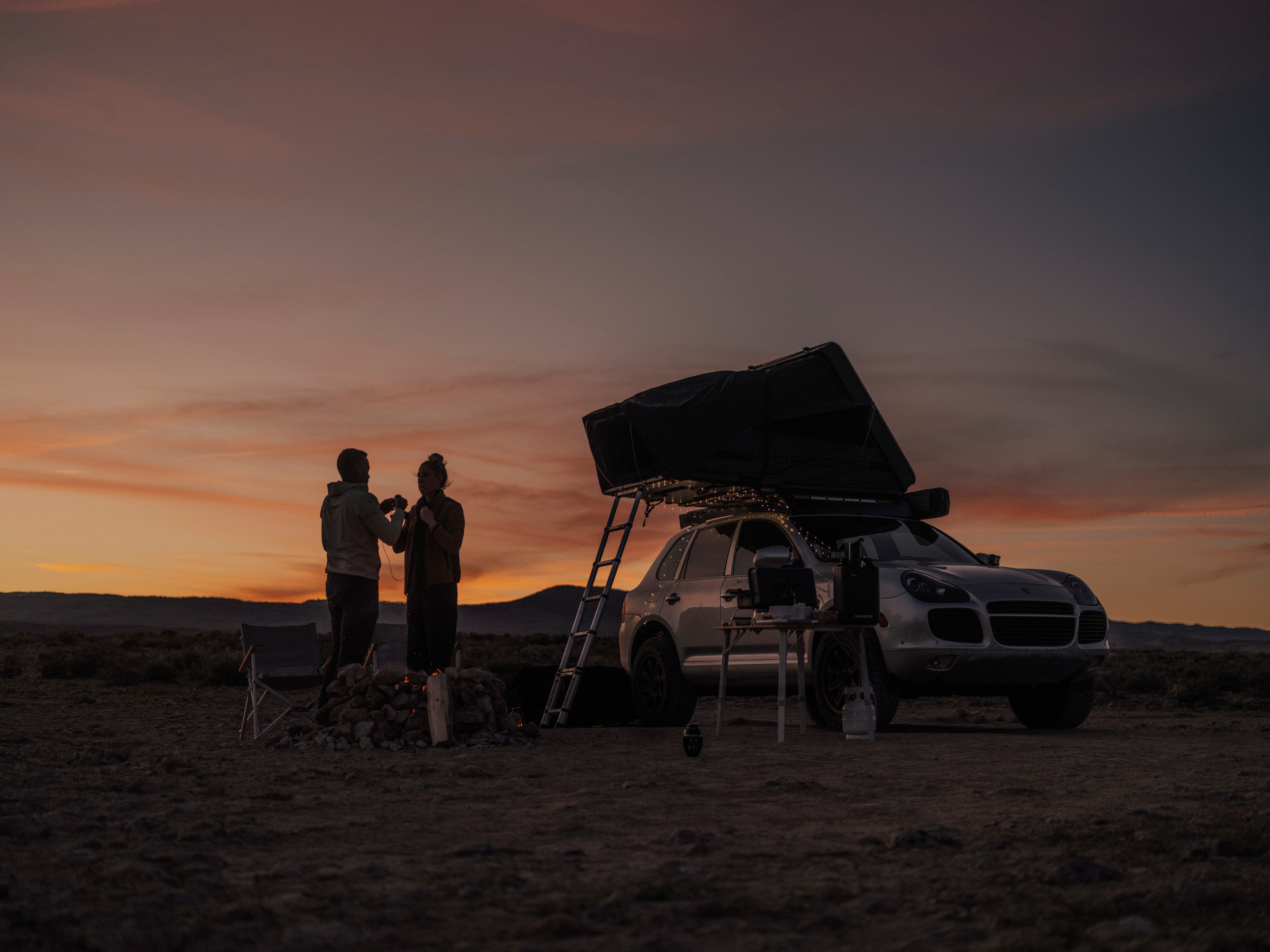 Couple standing beside Porsche Cayenne with roof-top tent as sun sets