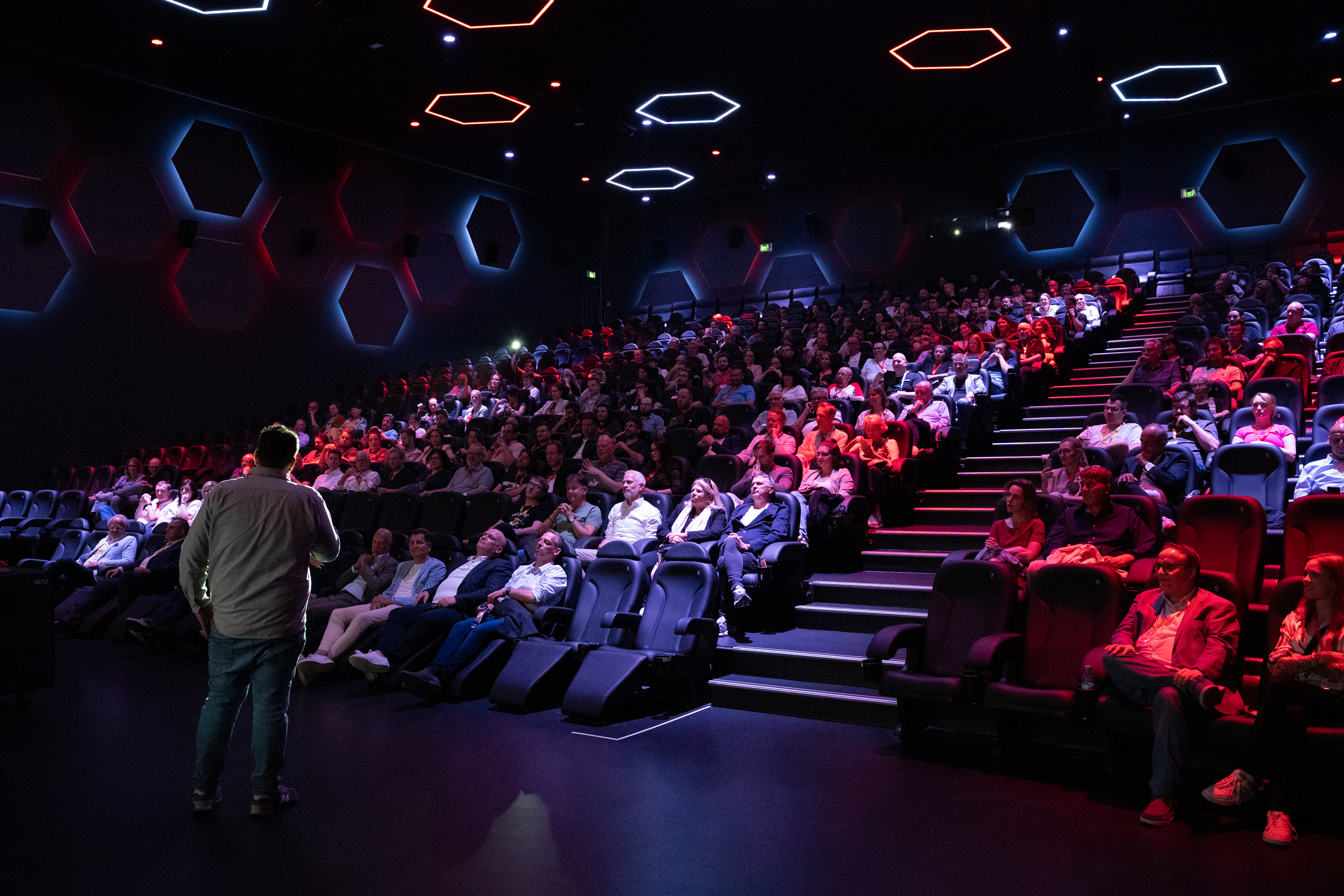 Hier sieht man einen vollen Kinosaal. Vor der Leinwand steht jemand, der eine Präsentation hält. 