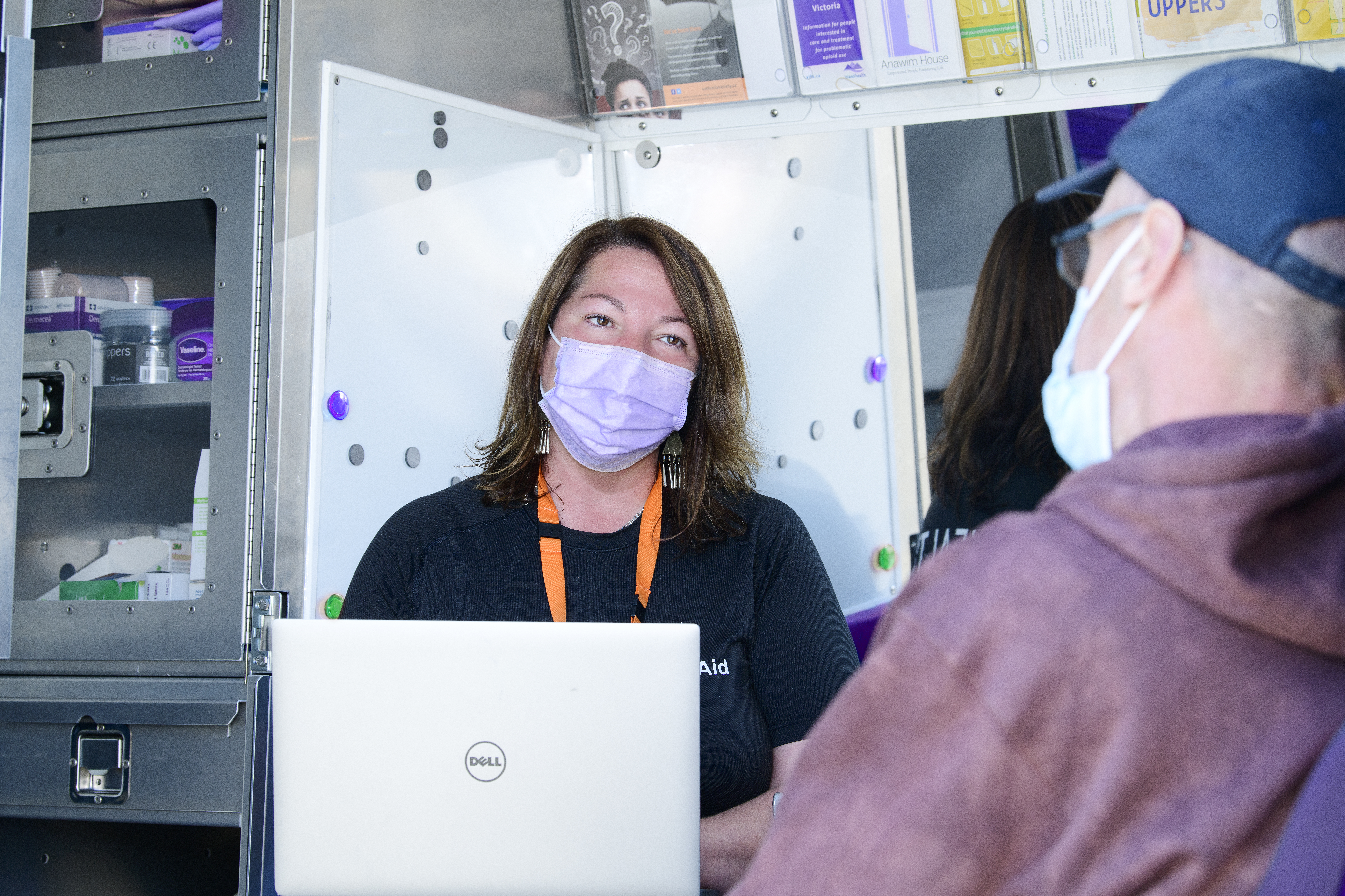 Candide Dias, Coordinator of Outreach Health Services at Cool Aid sitting in the Mobile Health Clinic van seeing a patient. Both Candide and the patient are wearing masks and Candide is on a laptop