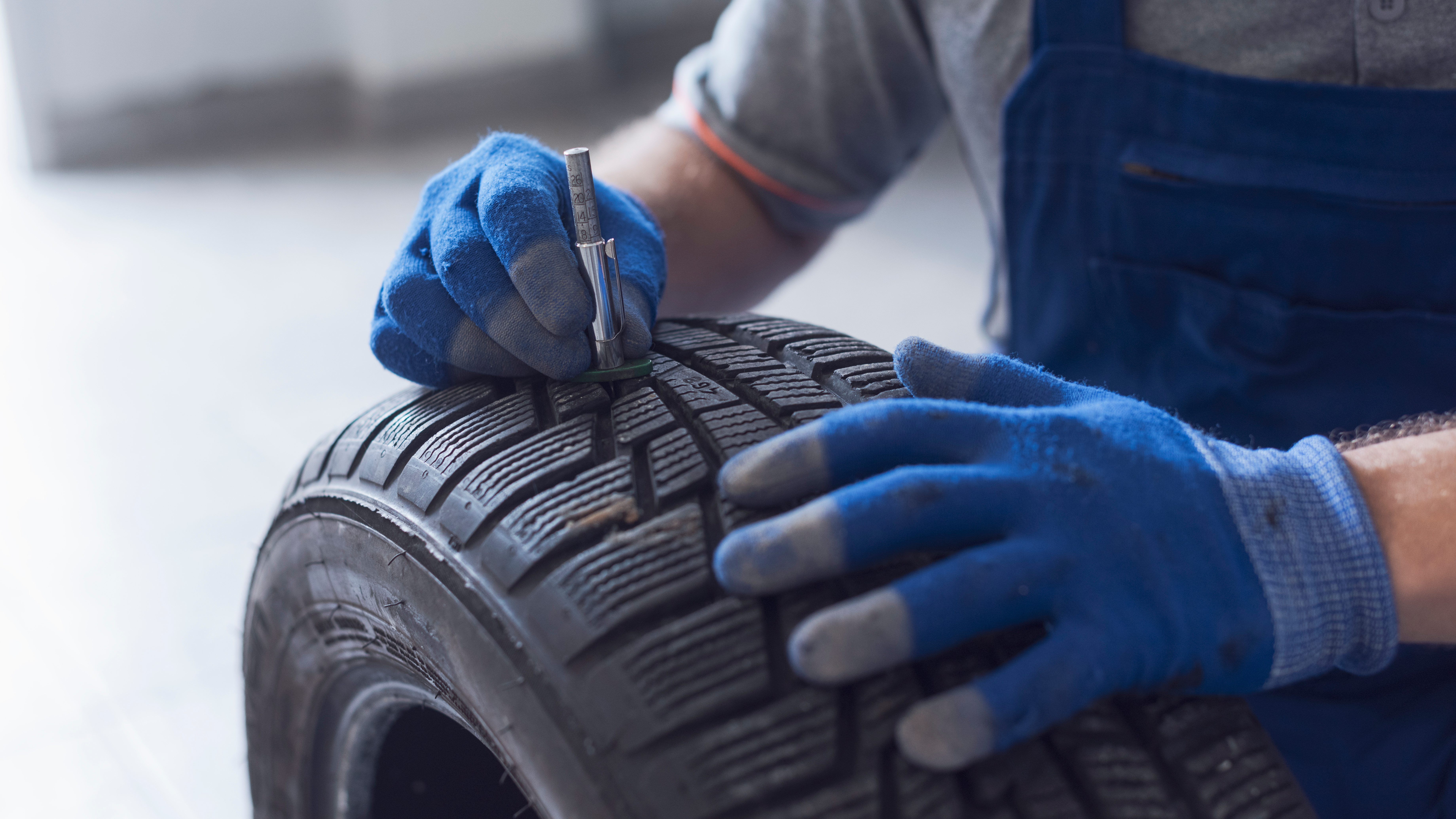 Nahaufnahme der Hände eines Mechanikers mit blauen Handschuhen. Er misst mit einem Profiltiefenmesser die Rillen eines Autoreifens.