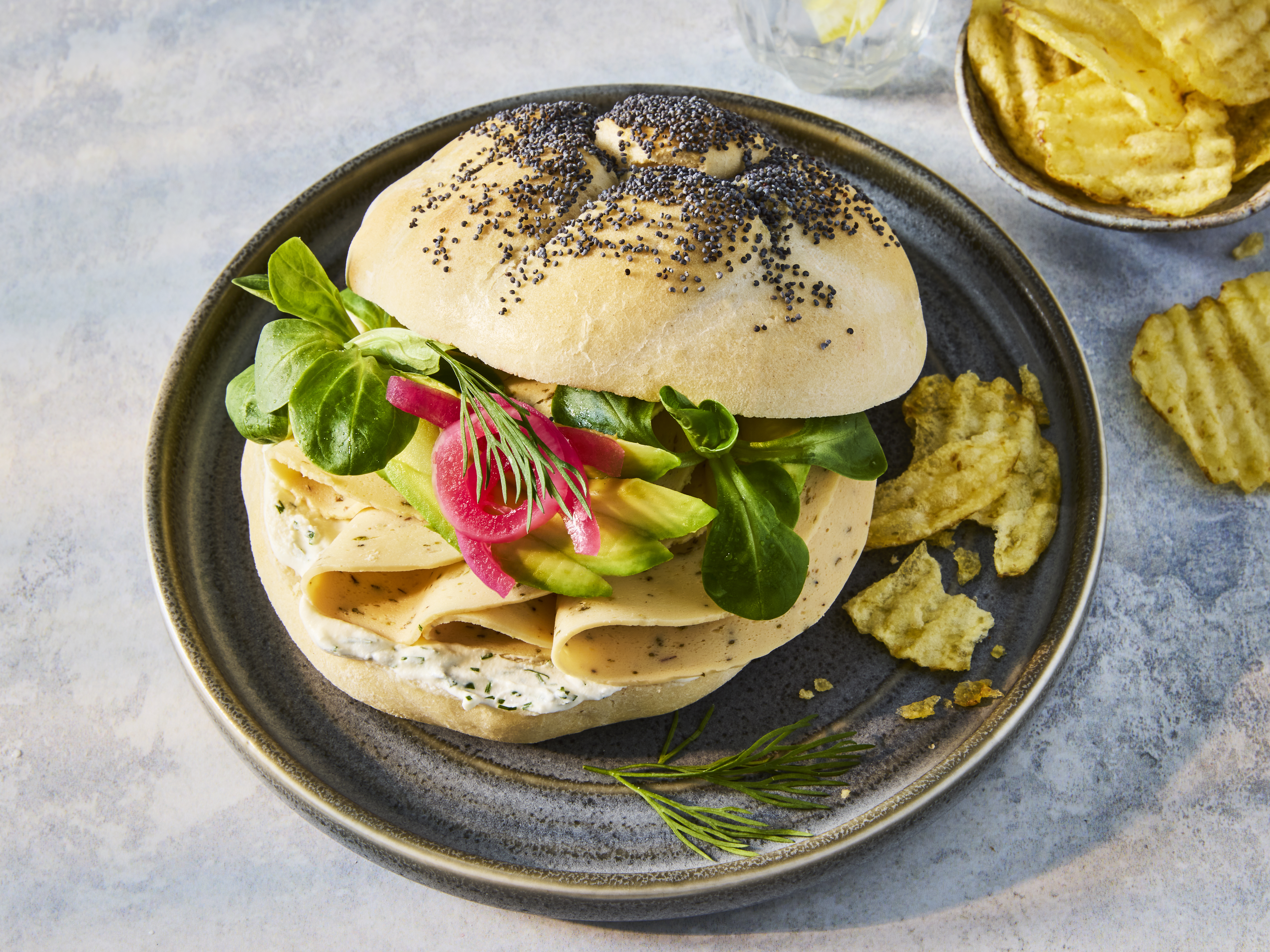 Sandwich on a navy plate with crisps on the side. Sandwich is made up of a white roll with black poppy seeds, Quorn Garlic & Herb Slices, salad, avocado and crème fraîche.