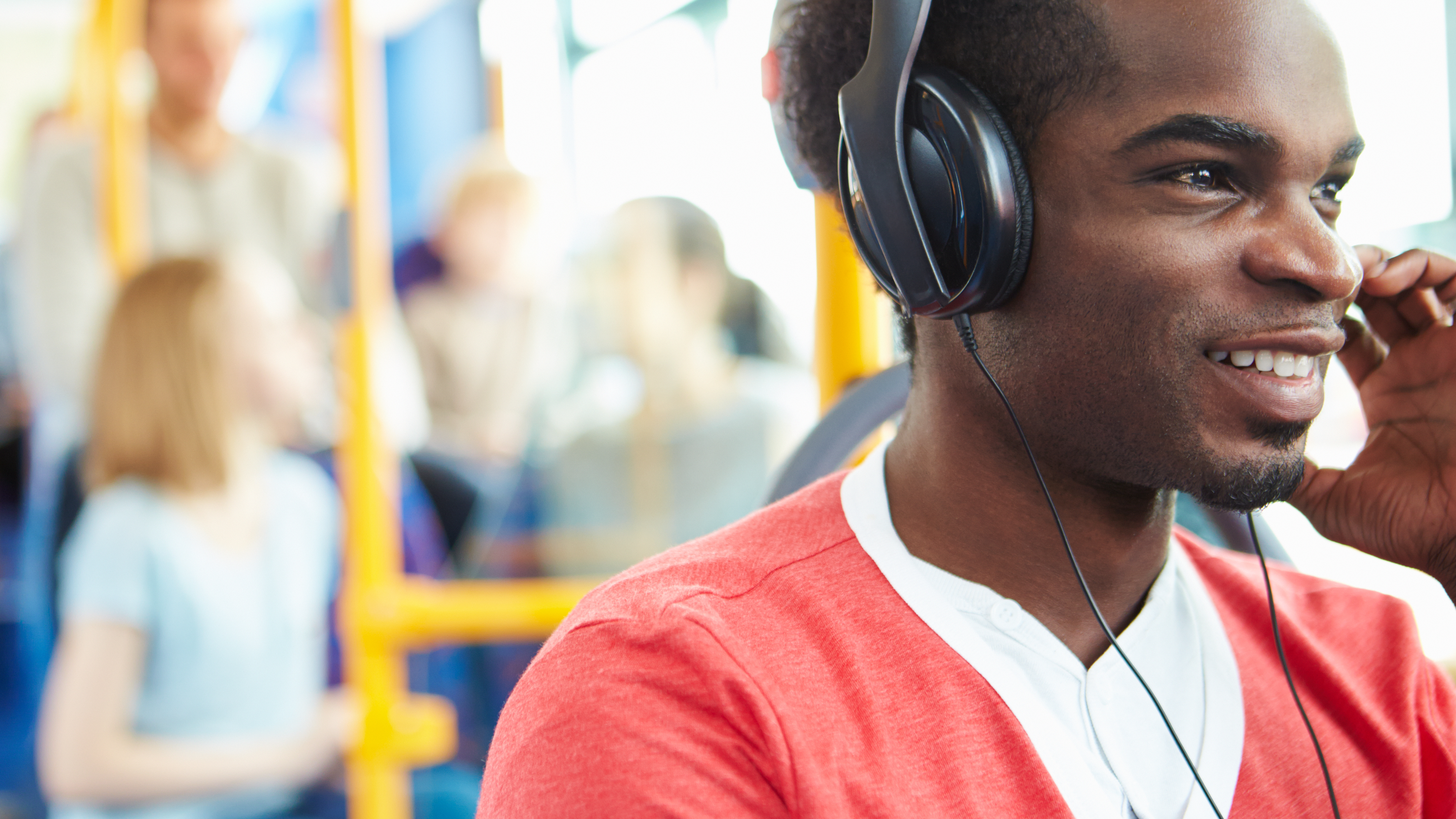 A male student wearing a red and white t-shirt and headphones sits on the bus listening to a podcast