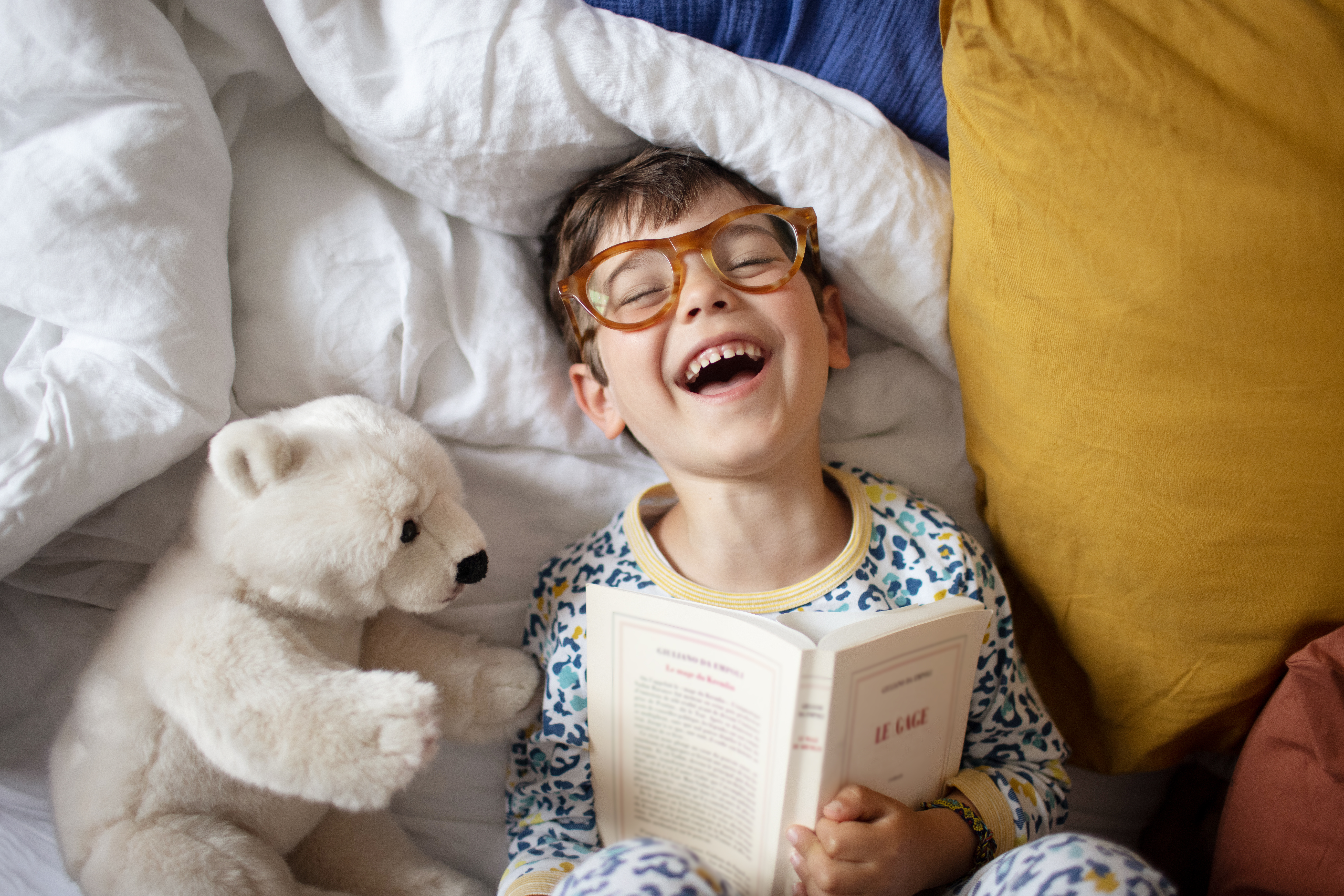 A young child wearing glasses and pajamas lies in bed, laughing while holding a book. A white stuffed bear is beside them on the bed, which is covered with white and yellow pillows and blankets.