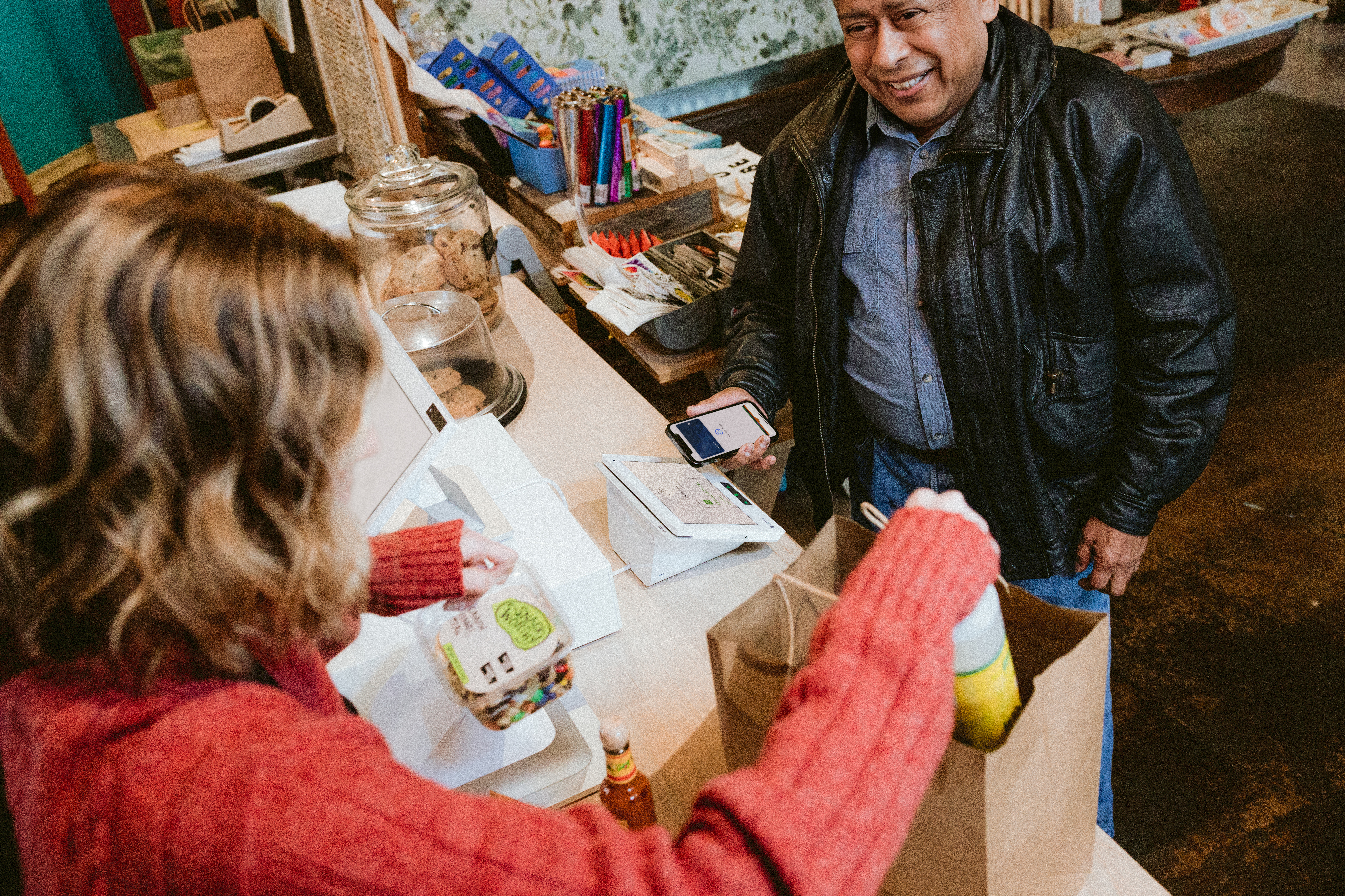 Customer buying groceries using tap to pay while cashier places groceries in bag.