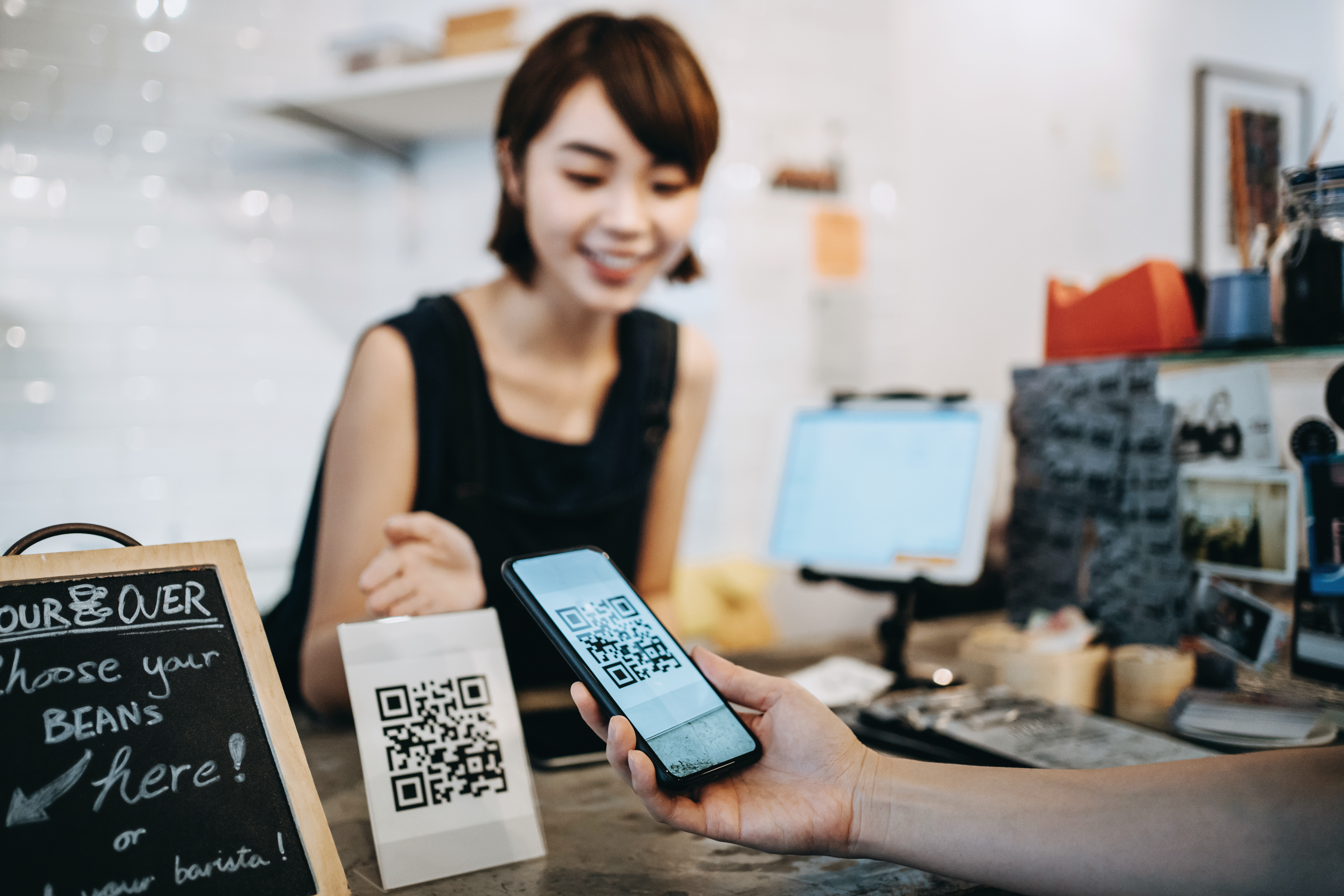 Barista in a coffee shop working with a customer to scan a QR code on a menu. Customer uses their phone to scan QR code. 