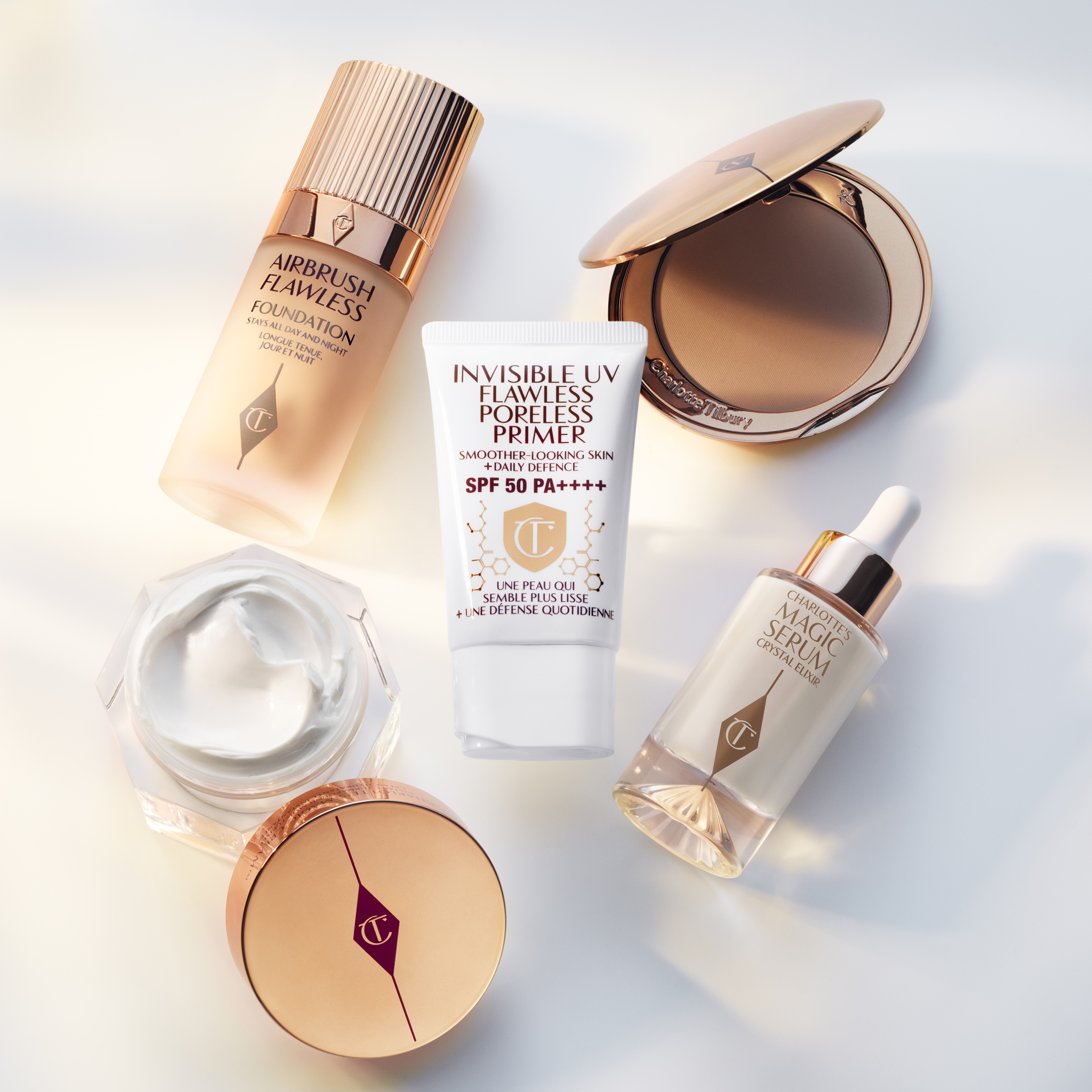 Foundation in a frosted glass bottle with a gold-coloured lid, bronzer compact in sleek, gold-coloured packaging, pearly-white face cream in a glass jar with a gold-coloured lid, luminous, ivory-coloured serum with a dropper lid, and a UV primer in a whit-coloured tube.