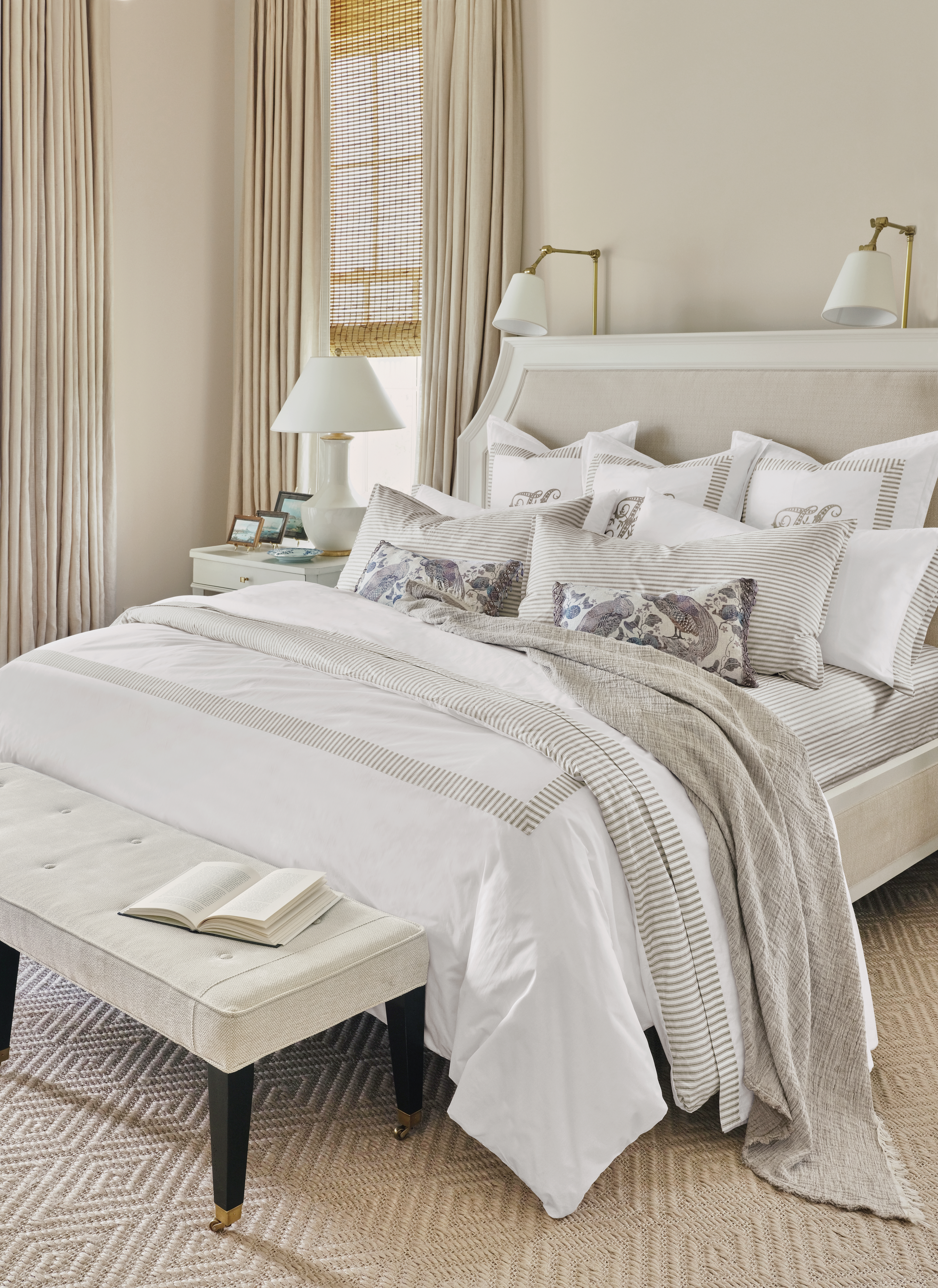 Bright neutral bedroom with upholstered bed, layered striped bedding, patterned accent pillows, beige throw blanket, white nightstand with lamp, woven blinds, and tufted bench holding an open book.