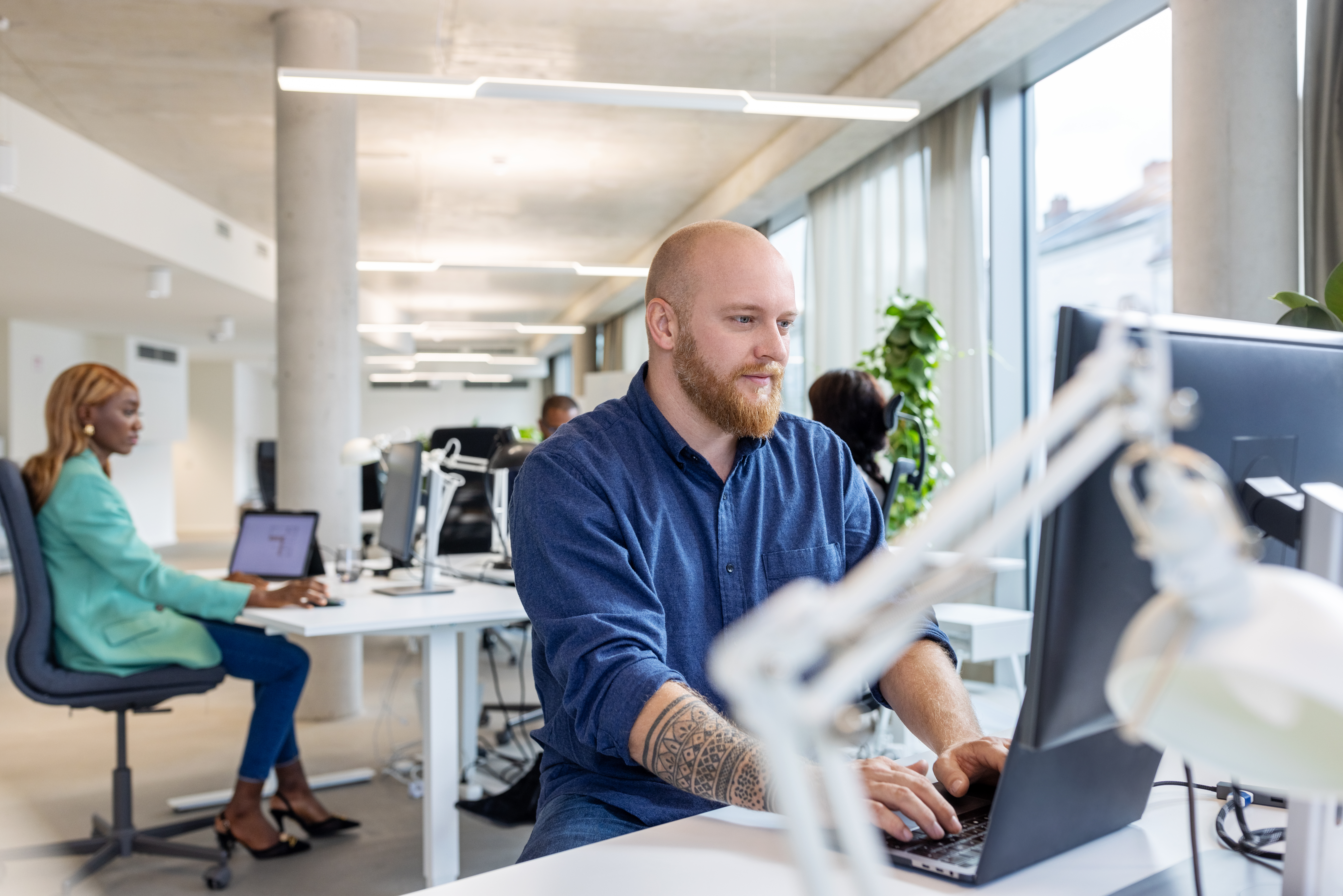 [Featured image] An IT support specialist in a blue shirt stands in front of a laptop in a brightly lit office. A co-worker sits in the background.