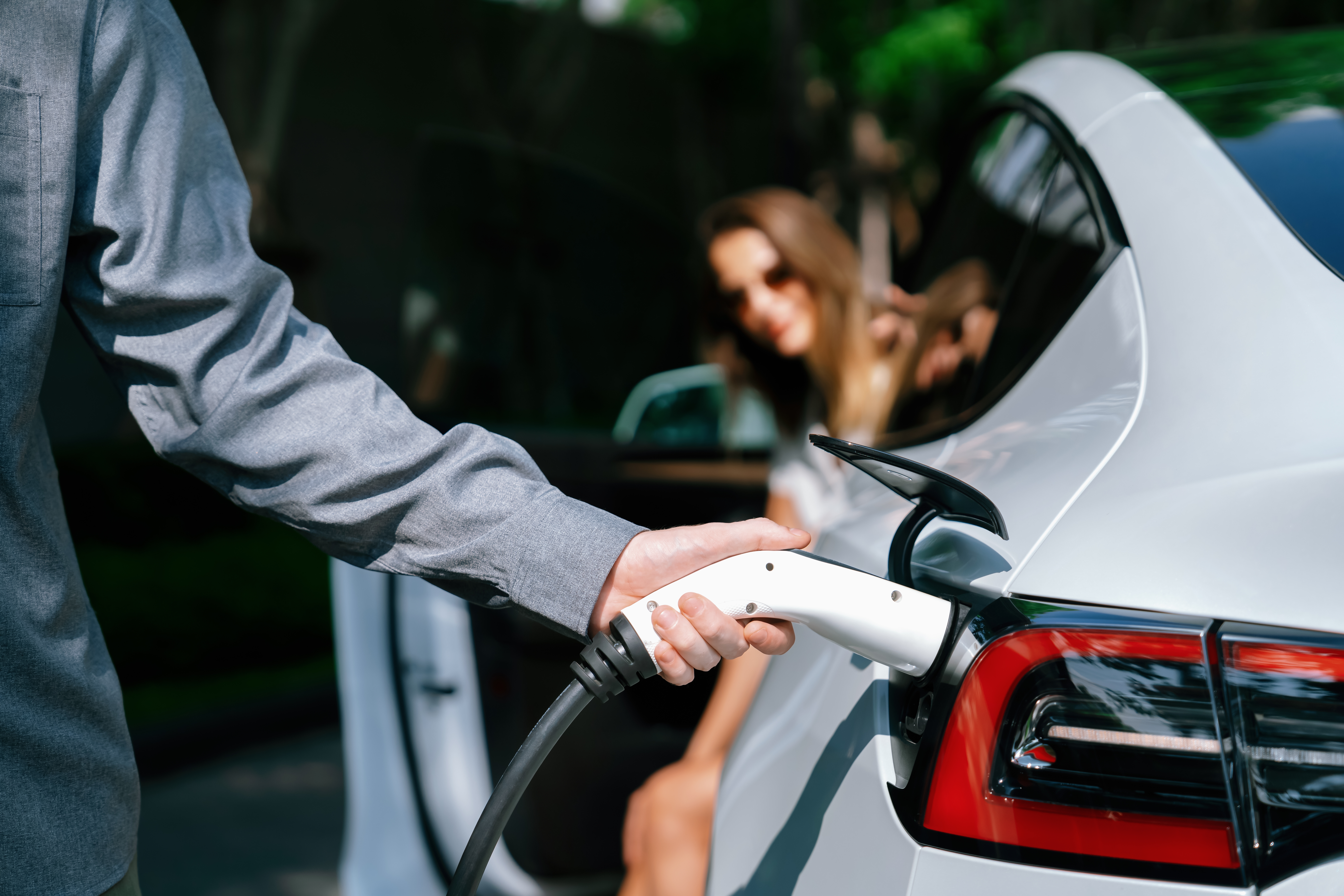 Personne rechargeant une voiture électrique tandis qu'une femme sourit à l'arrière du véhicule.