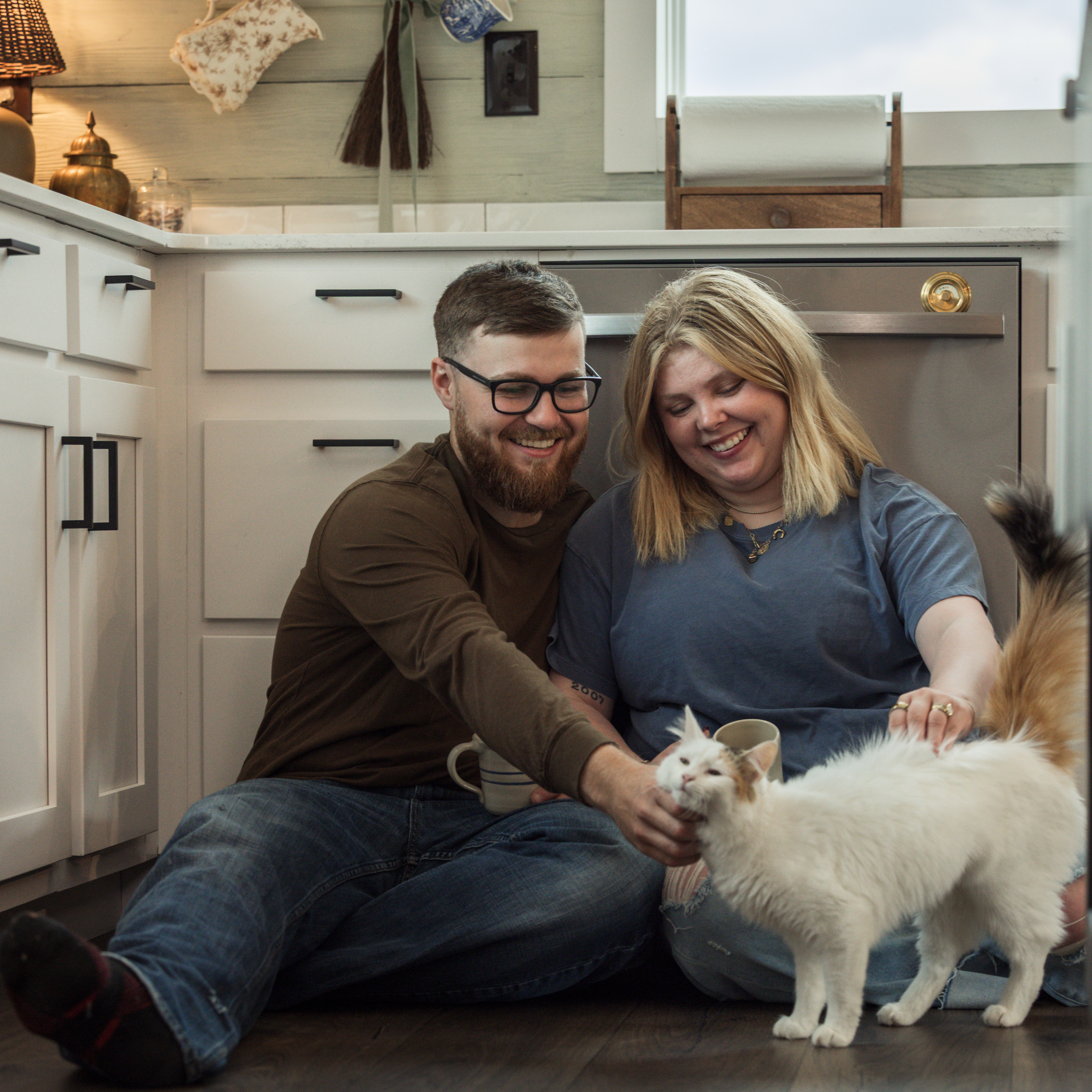 A young couple smile and sit against their kitchen cabinets on the floor while petting their white and orange cat.