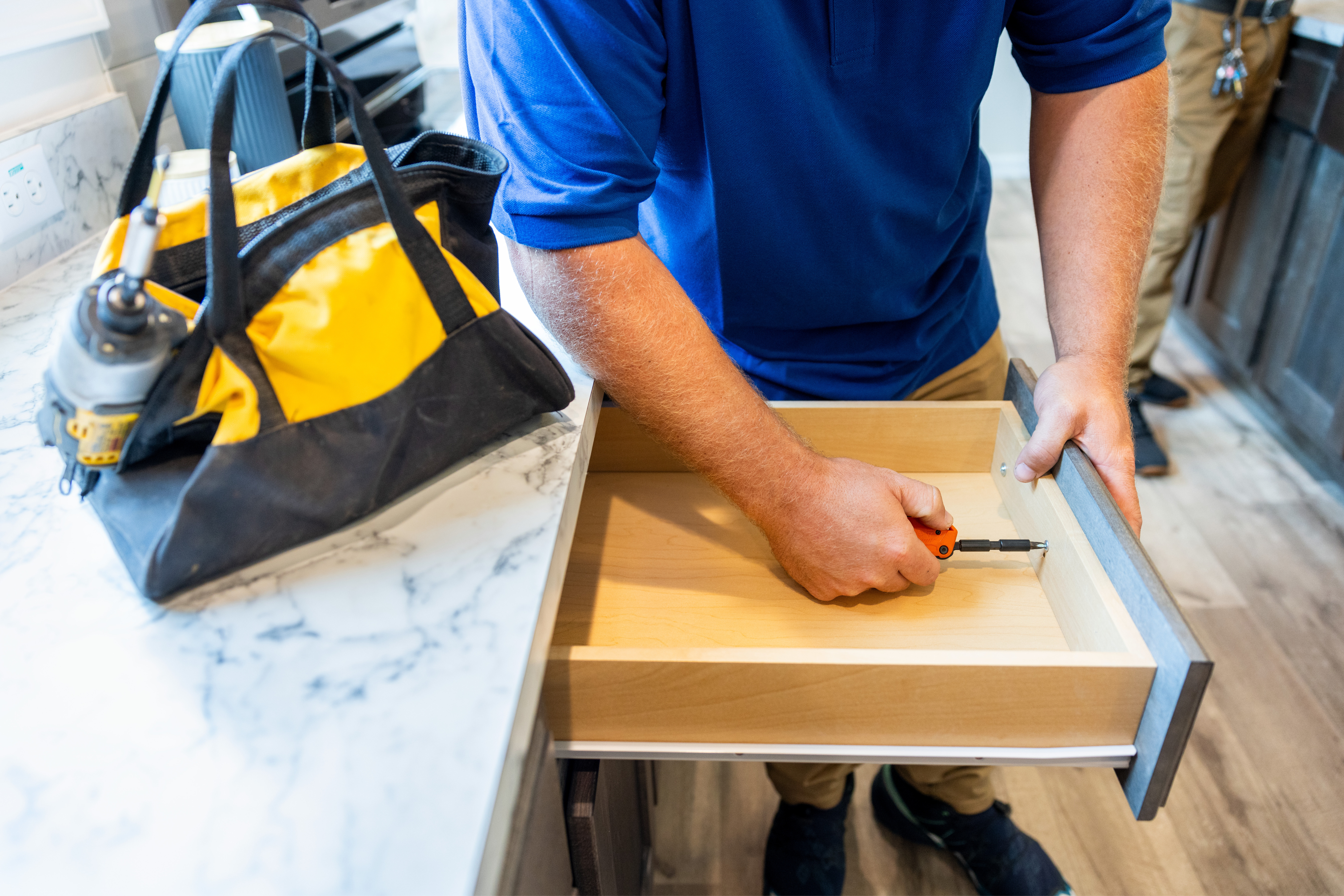 A service worker from the shoulders down uses a tool to work on a kitchen drawer with his yellow tool bag on the counter.