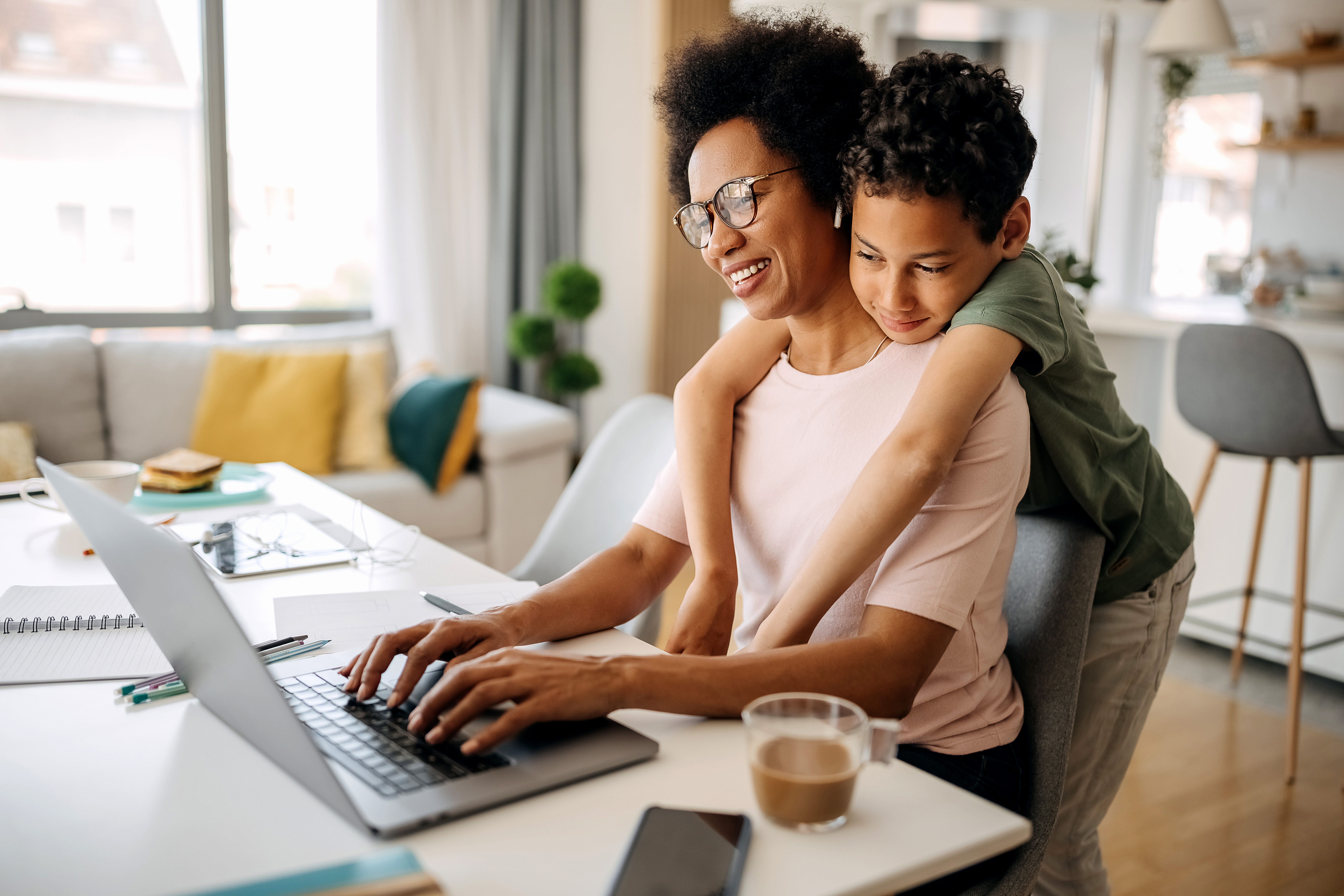 Mother smiles as she works on her laptop on the kitchen table while her son snuggles her from behind